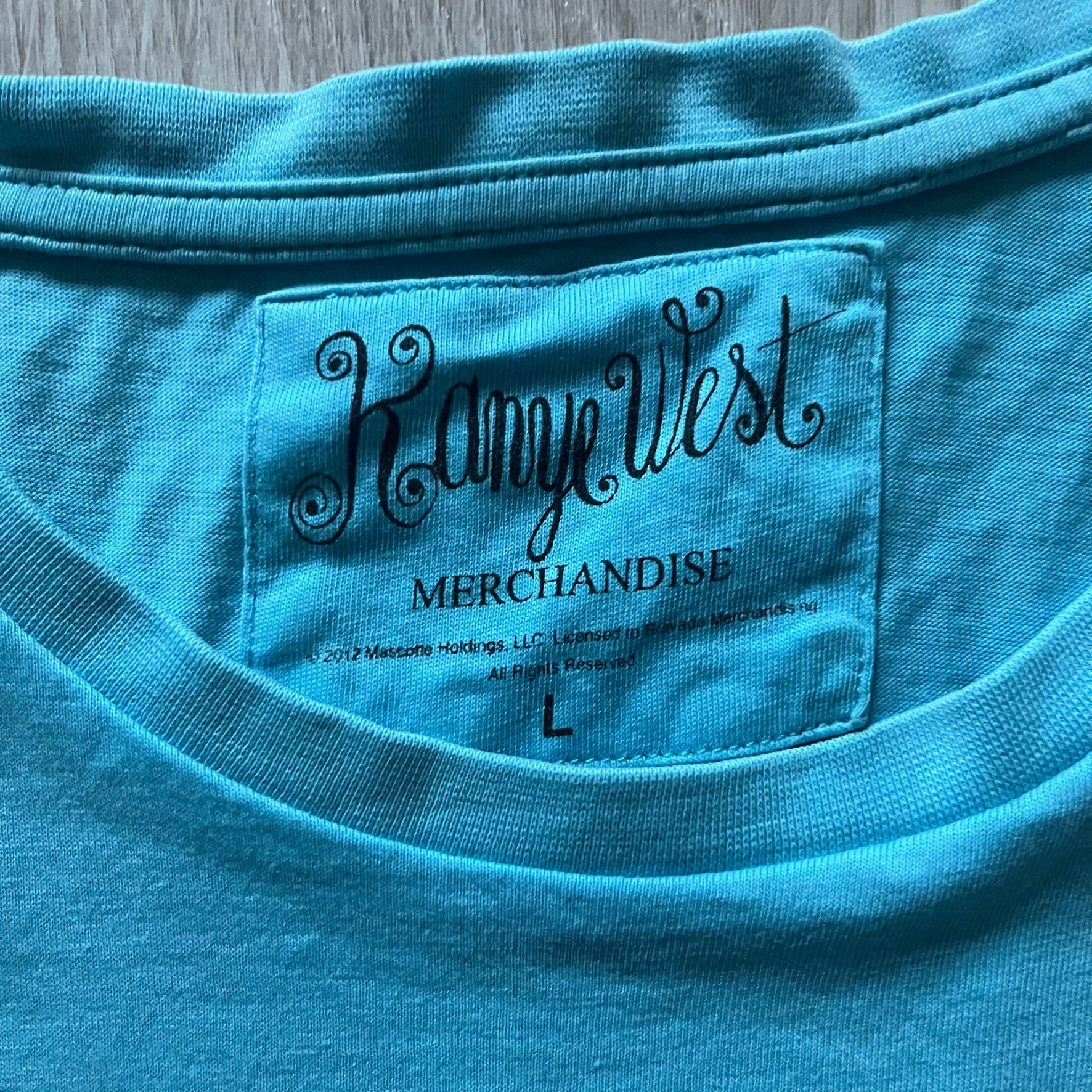 Kanye West 2012 Takashi Murakami 'Glow In The Dark Tour' Tee in light blue features the iconic bear with shutter glasses on the front. Below the graphic is yellow "kanYeWest" text and the 2007 Takashi Murakami/Kanye West licensing. Official merchandise from the 2012, this collectible tee includes the neck tag with Kanye logo and Mascotte Holdings licensing.