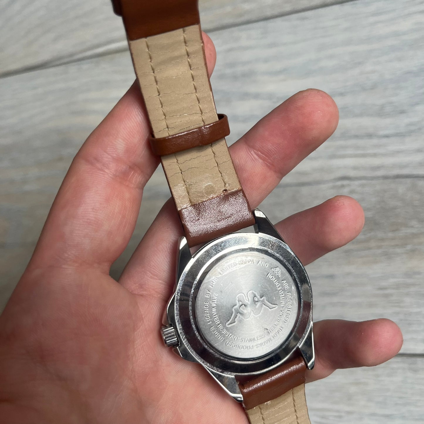 Kappa 2000s Quartz Analog Watch features brown leather straps, a silver-tone case, and a white dial. The bezel is marked in five-minute increments from 5 to 55. The dial has a Kappa logo at the top and Kappa text below the center. The hour markers are represented by white dots and numerals. The watch has a stainless steel case back and is marked as a limited edition model.