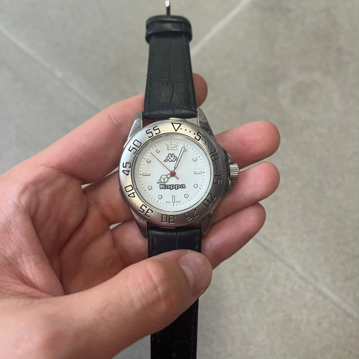 Kappa 2000's Quartz Analog Watch features black leather straps, a silver-tone case, and a white dial. The bezel is marked in five-minute increments from 5 to 55. The dial has a Kappa logo at the top and Kappa text below the center. The hour markers are represented by white dots and numerals. The watch has a stainless steel case back and is marked as a limited edition model.