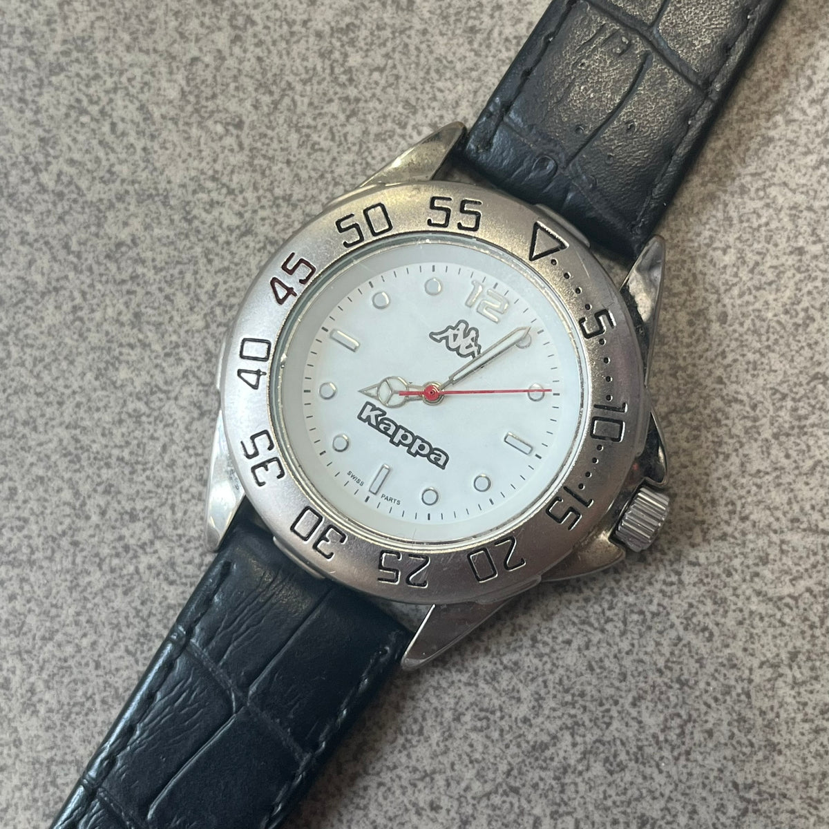 Kappa 2000's Quartz Analog Watch features black leather straps, a silver-tone case, and a white dial. The bezel is marked in five-minute increments from 5 to 55. The dial has a Kappa logo at the top and Kappa text below the center. The hour markers are represented by white dots and numerals. The watch has a stainless steel case back and is marked as a limited edition model.