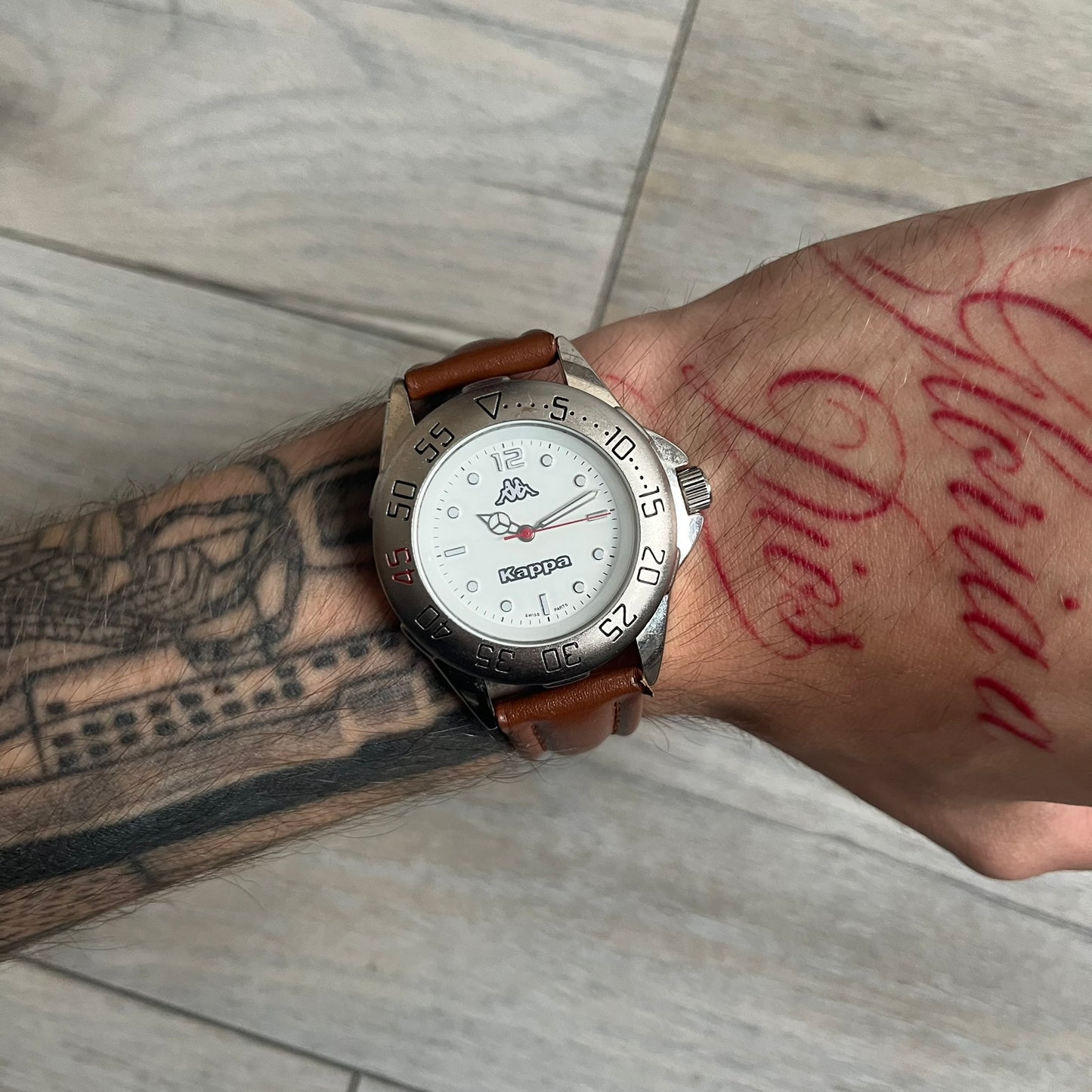Kappa 2000s Quartz Analog Watch features brown leather straps, a silver-tone case, and a white dial. The bezel is marked in five-minute increments from 5 to 55. The dial has a Kappa logo at the top and Kappa text below the center. The hour markers are represented by white dots and numerals. The watch has a stainless steel case back and is marked as a limited edition model.
