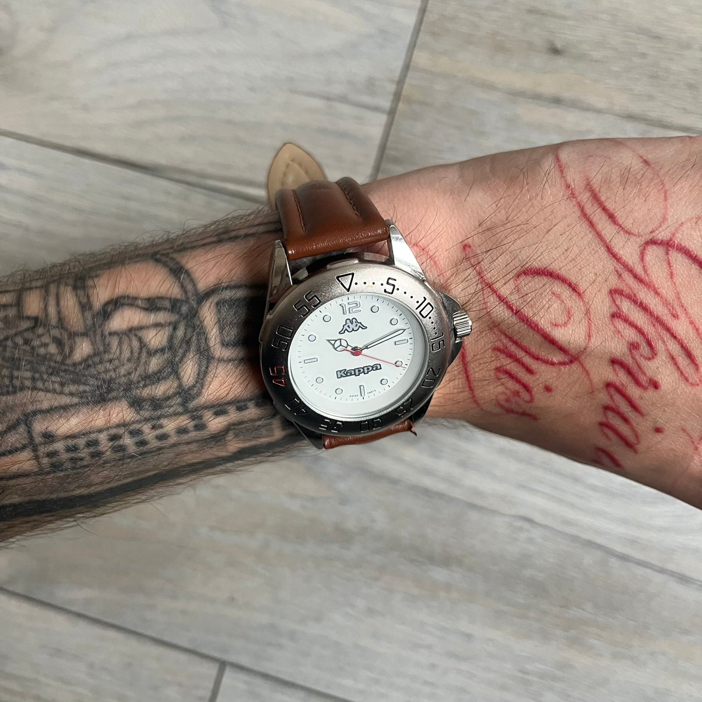 Kappa 2000s Quartz Analog Watch features brown leather straps, a silver-tone case, and a white dial. The bezel is marked in five-minute increments from 5 to 55. The dial has a Kappa logo at the top and Kappa text below the center. The hour markers are represented by white dots and numerals. The watch has a stainless steel case back and is marked as a limited edition model.