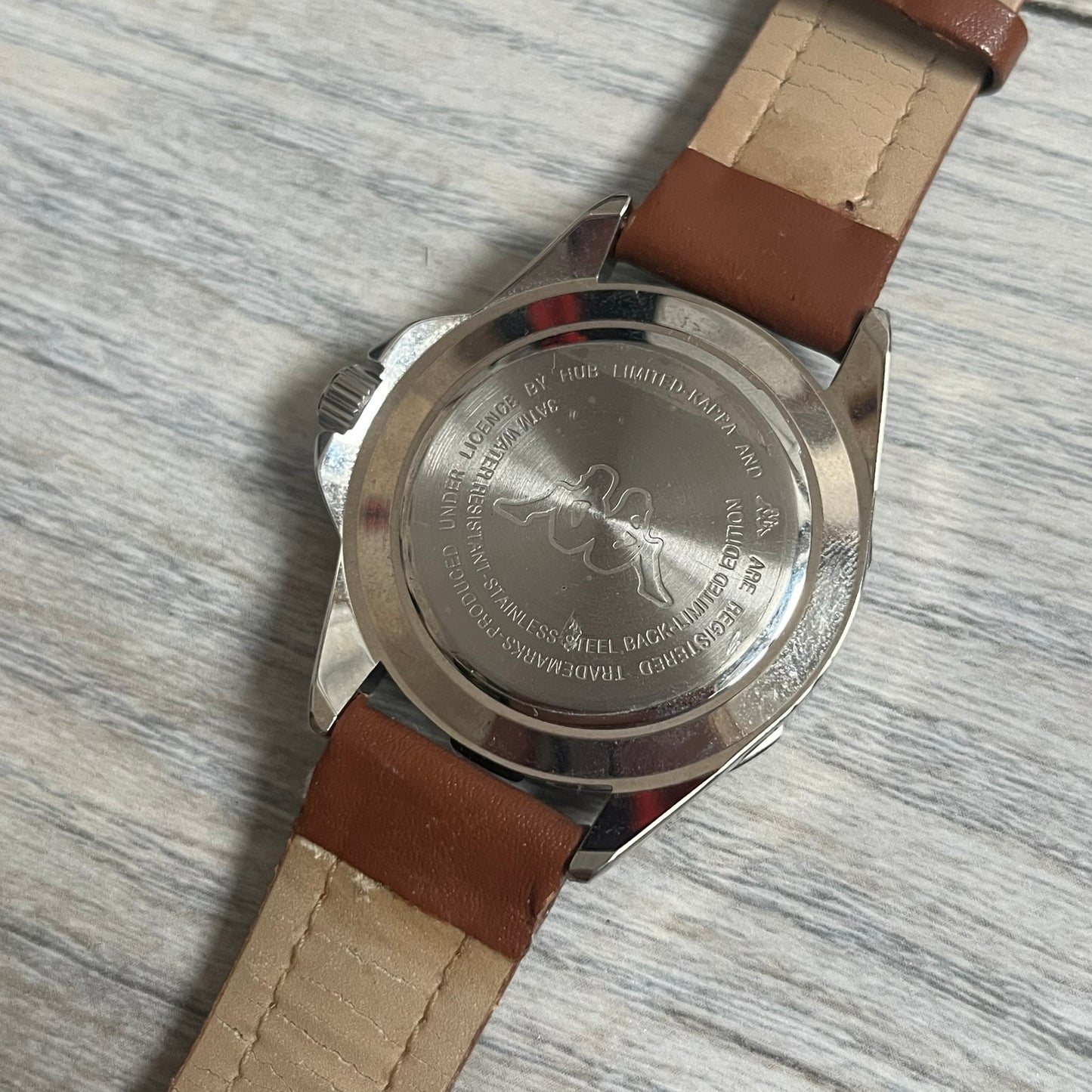 Kappa 2000s Quartz Analog Watch features brown leather straps, a silver-tone case, and a white dial. The bezel is marked in five-minute increments from 5 to 55. The dial has a Kappa logo at the top and Kappa text below the center. The hour markers are represented by white dots and numerals. The watch has a stainless steel case back and is marked as a limited edition model.