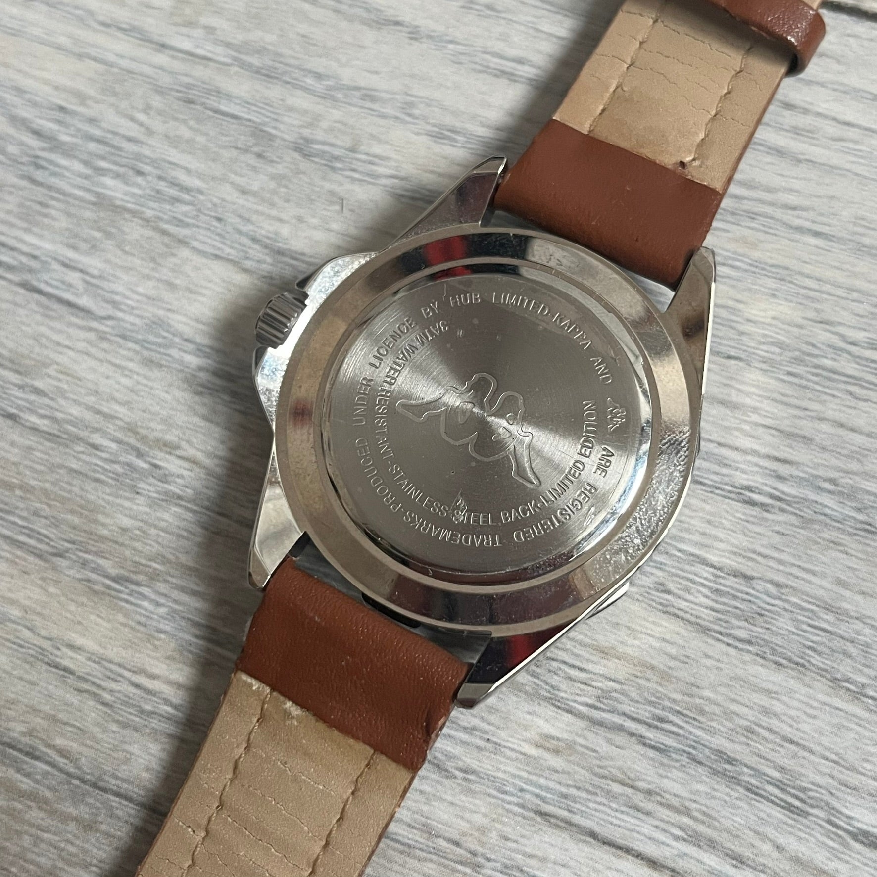Kappa 2000s Quartz Analog Watch features brown leather straps, a silver-tone case, and a white dial. The bezel is marked in five-minute increments from 5 to 55. The dial has a Kappa logo at the top and Kappa text below the center. The hour markers are represented by white dots and numerals. The watch has a stainless steel case back and is marked as a limited edition model.