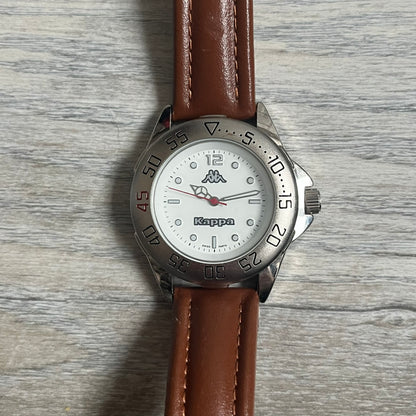 Kappa 2000s Quartz Analog Watch features brown leather straps, a silver-tone case, and a white dial. The bezel is marked in five-minute increments from 5 to 55. The dial has a Kappa logo at the top and Kappa text below the center. The hour markers are represented by white dots and numerals. The watch has a stainless steel case back and is marked as a limited edition model.