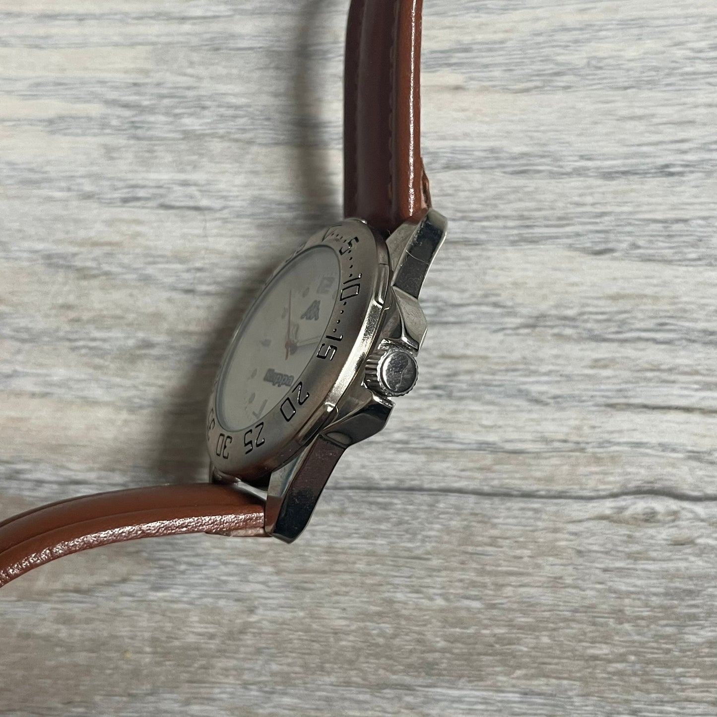 Kappa 2000s Quartz Analog Watch features brown leather straps, a silver-tone case, and a white dial. The bezel is marked in five-minute increments from 5 to 55. The dial has a Kappa logo at the top and Kappa text below the center. The hour markers are represented by white dots and numerals. The watch has a stainless steel case back and is marked as a limited edition model.