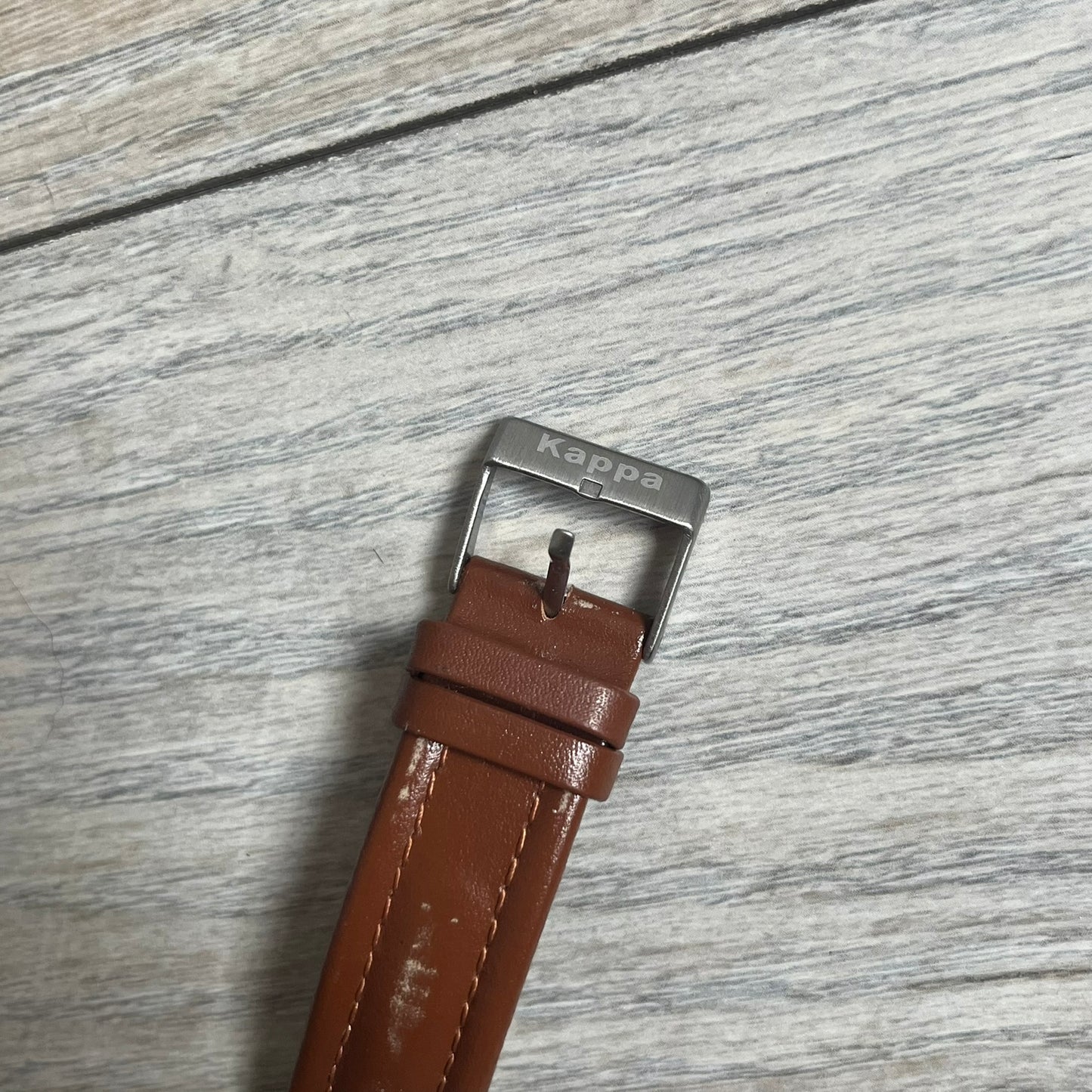 Kappa 2000s Quartz Analog Watch features brown leather straps, a silver-tone case, and a white dial. The bezel is marked in five-minute increments from 5 to 55. The dial has a Kappa logo at the top and Kappa text below the center. The hour markers are represented by white dots and numerals. The watch has a stainless steel case back and is marked as a limited edition model.