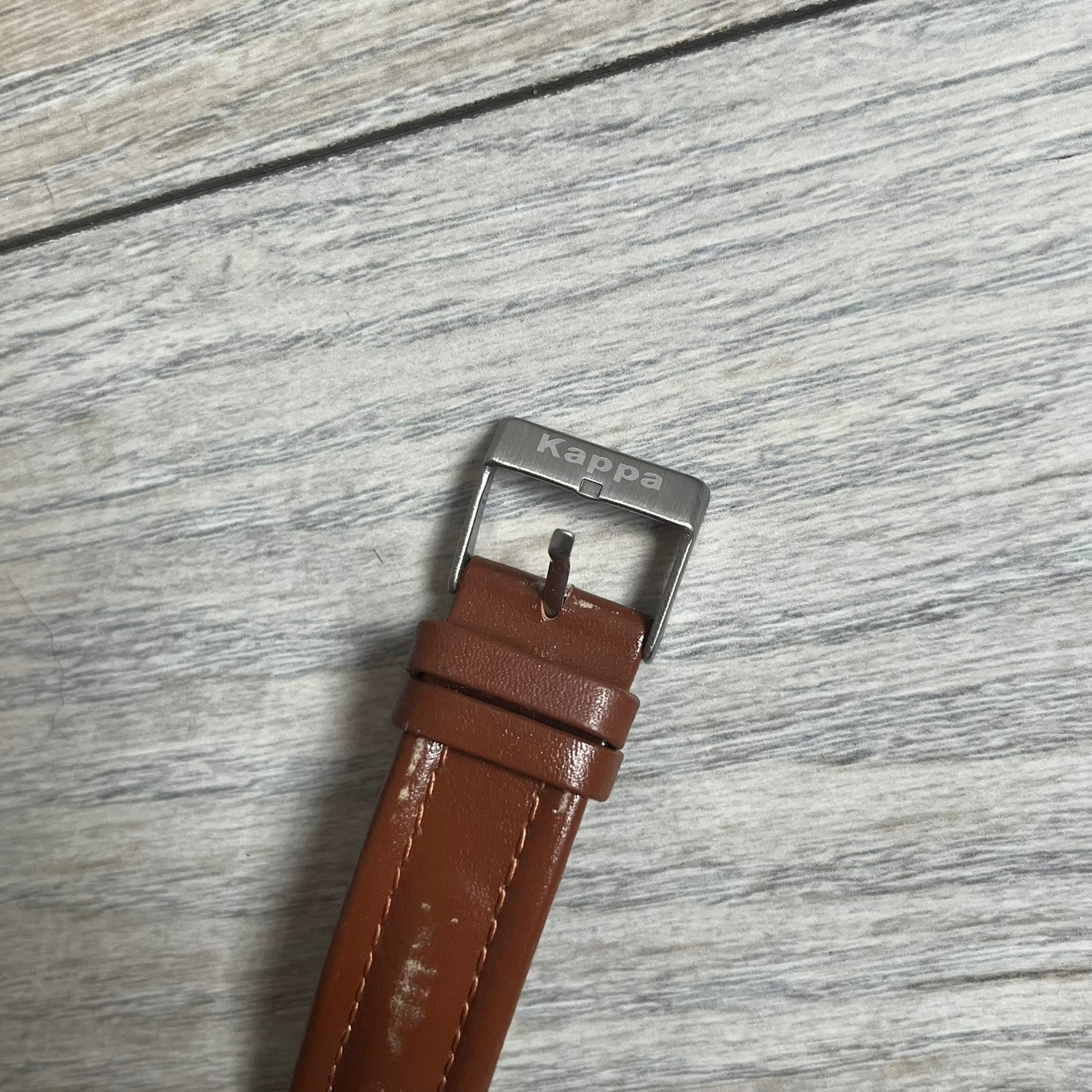Kappa 2000s Quartz Analog Watch features brown leather straps, a silver-tone case, and a white dial. The bezel is marked in five-minute increments from 5 to 55. The dial has a Kappa logo at the top and Kappa text below the center. The hour markers are represented by white dots and numerals. The watch has a stainless steel case back and is marked as a limited edition model.