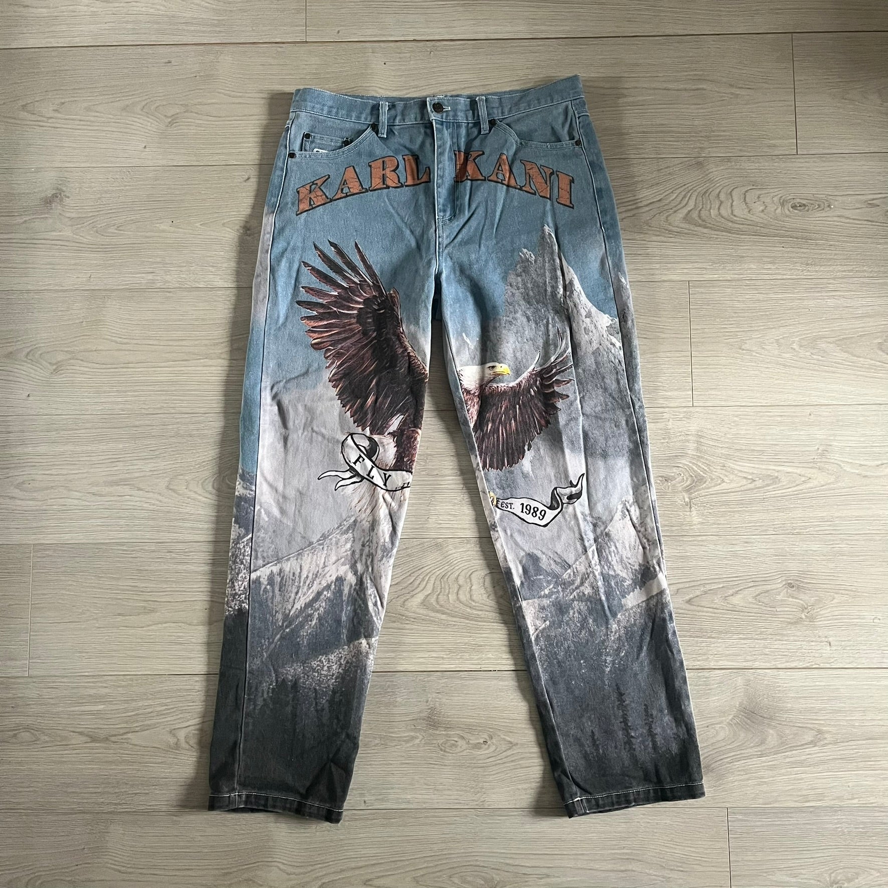 The Karl Kani Signature Eagle Printed Jeans feature a multi-tone eagle graphic over a mountain landscape on a classic blue denim base. Made from 100% cotton, these baggy jeans have a relaxed, wide-leg fit with straight cut legs, a button and zip fastening, and embroidered Karl Kani branding on the back and front pockets. The all-over print extends across the front and back, creating a unique and vintage-inspired look from the FW22 Season.