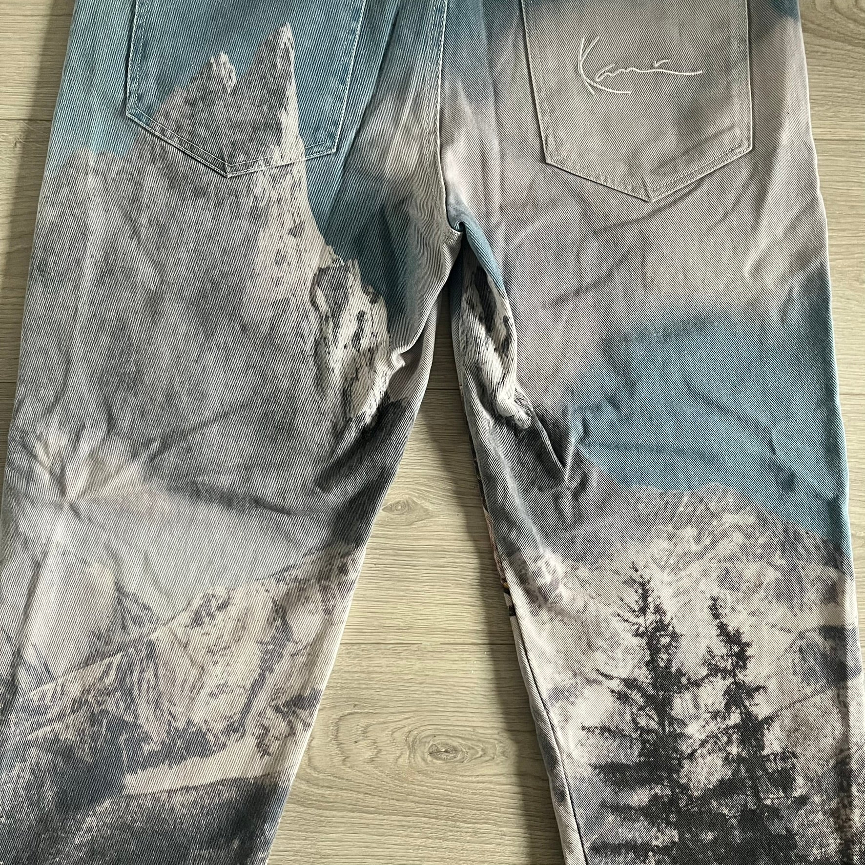 The Karl Kani Signature Eagle Printed Jeans feature a multi-tone eagle graphic over a mountain landscape on a classic blue denim base. Made from 100% cotton, these baggy jeans have a relaxed, wide-leg fit with straight cut legs, a button and zip fastening, and embroidered Karl Kani branding on the back and front pockets. The all-over print extends across the front and back, creating a unique and vintage-inspired look from the FW22 Season.