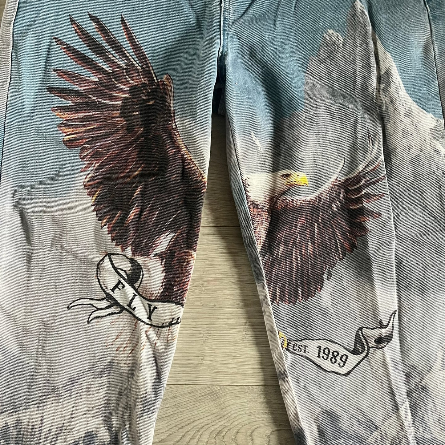 The Karl Kani Signature Eagle Printed Jeans feature a multi-tone eagle graphic over a mountain landscape on a classic blue denim base. Made from 100% cotton, these baggy jeans have a relaxed, wide-leg fit with straight cut legs, a button and zip fastening, and embroidered Karl Kani branding on the back and front pockets. The all-over print extends across the front and back, creating a unique and vintage-inspired look from the FW22 Season.