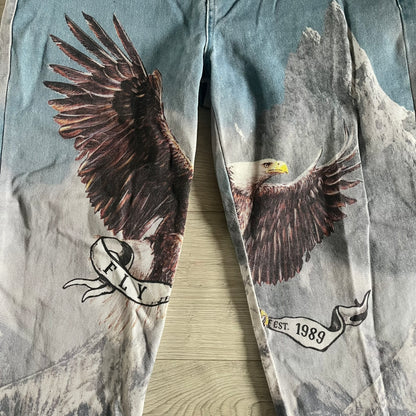 The Karl Kani Signature Eagle Printed Jeans feature a multi-tone eagle graphic over a mountain landscape on a classic blue denim base. Made from 100% cotton, these baggy jeans have a relaxed, wide-leg fit with straight cut legs, a button and zip fastening, and embroidered Karl Kani branding on the back and front pockets. The all-over print extends across the front and back, creating a unique and vintage-inspired look from the FW22 Season.
