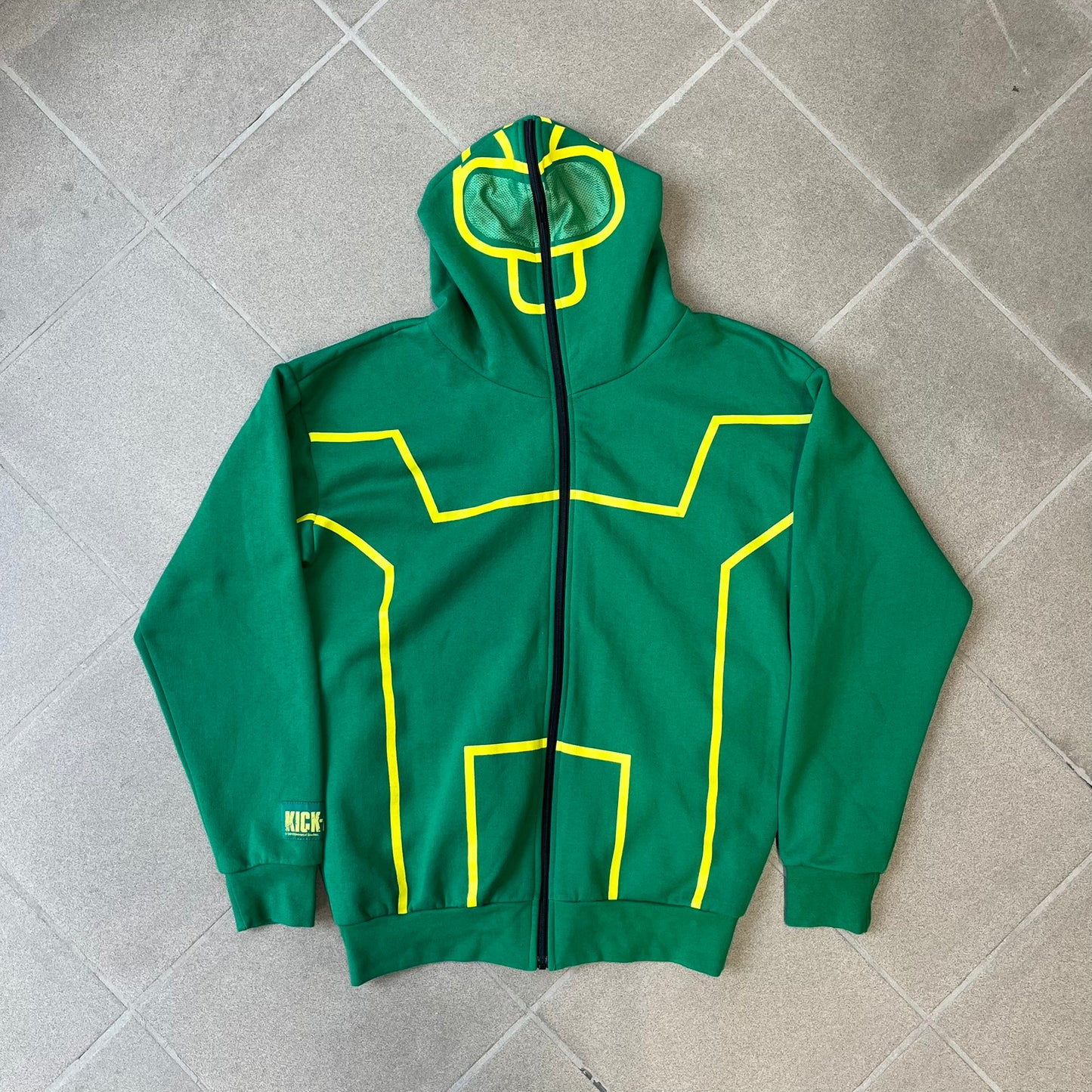 Kick-Ass 2 Full Zip 2013 Promo Hoodie is a green full-zip hoodie with yellow printed lines matching the costume worn by Kick-Ass in Kick-Ass 2. The zipper extends through the hood, allowing it to close into a mask with see-through mesh eye panels. It features a Kick-Ass 2 (2013) Universal Studios licensed neck tag, a matching movie logo patch on the sleeve, and is made in the USA.
