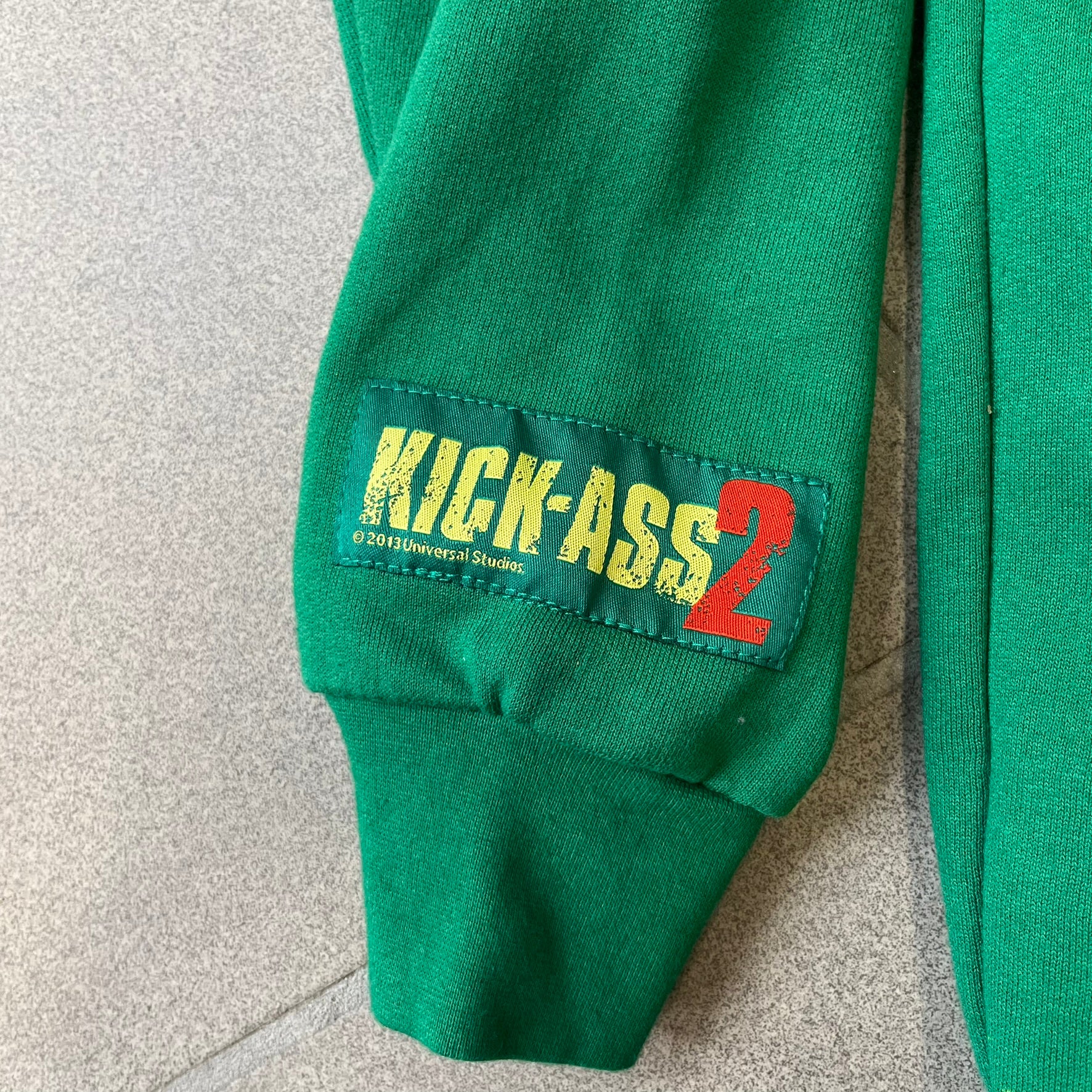 Kick-Ass 2 Full Zip 2013 Promo Hoodie is a green full-zip hoodie with yellow printed lines matching the costume worn by Kick-Ass in Kick-Ass 2. The zipper extends through the hood, allowing it to close into a mask with see-through mesh eye panels. It features a Kick-Ass 2 (2013) Universal Studios licensed neck tag, a matching movie logo patch on the sleeve, and is made in the USA.
