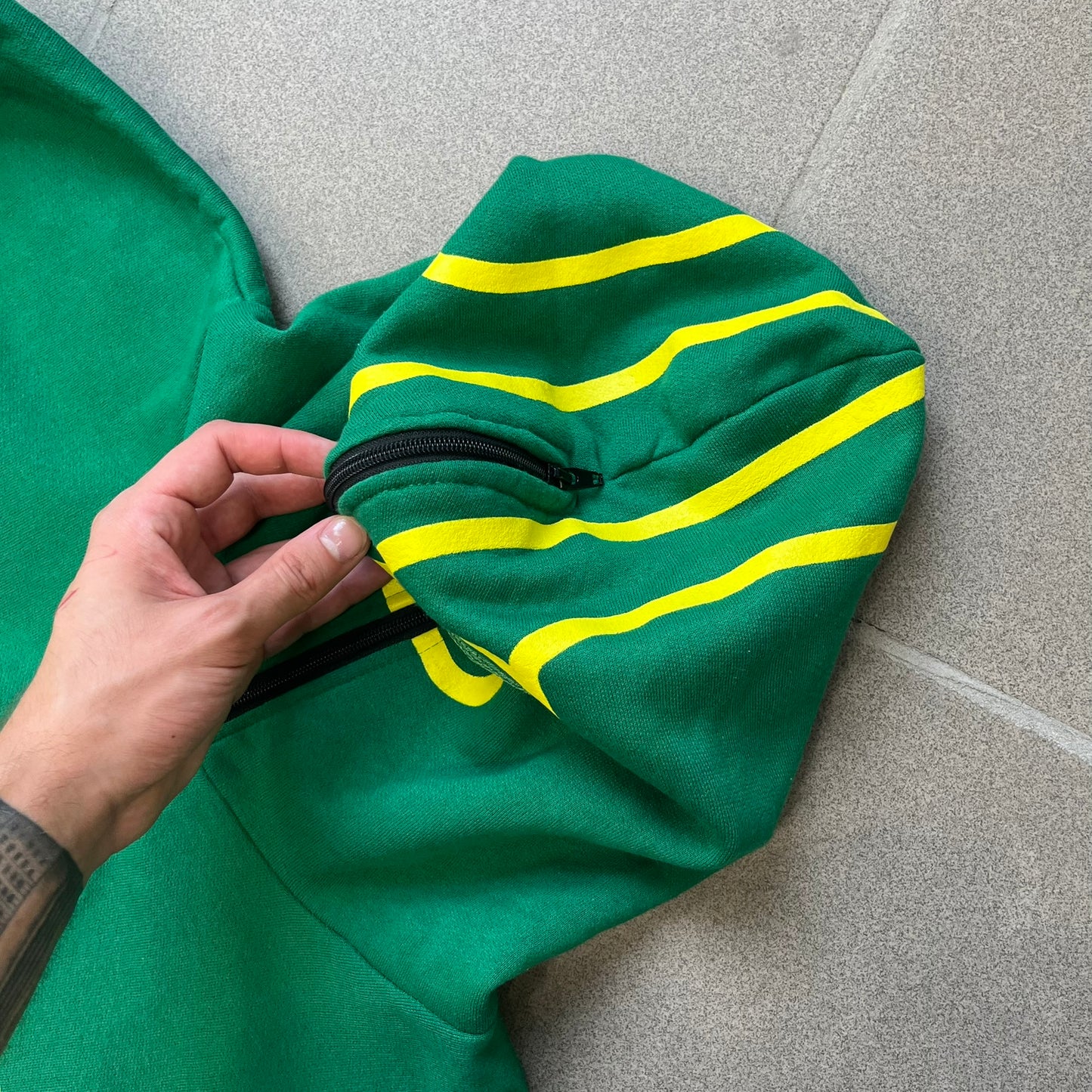 Kick-Ass 2 Full Zip 2013 Promo Hoodie is a green full-zip hoodie with yellow printed lines matching the costume worn by Kick-Ass in Kick-Ass 2. The zipper extends through the hood, allowing it to close into a mask with see-through mesh eye panels. It features a Kick-Ass 2 (2013) Universal Studios licensed neck tag, a matching movie logo patch on the sleeve, and is made in the USA.
