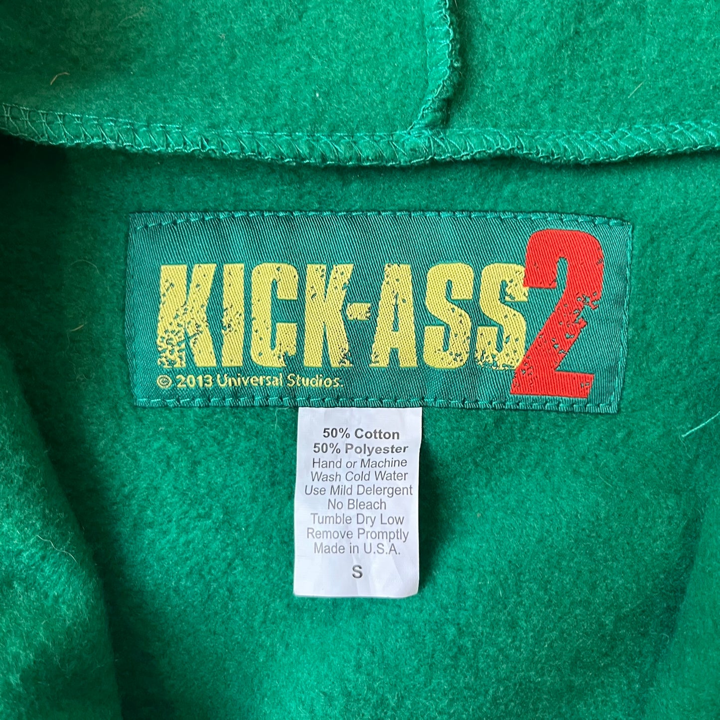 Kick-Ass 2 Full Zip 2013 Promo Hoodie is a green full-zip hoodie with yellow printed lines matching the costume worn by Kick-Ass in Kick-Ass 2. The zipper extends through the hood, allowing it to close into a mask with see-through mesh eye panels. It features a Kick-Ass 2 (2013) Universal Studios licensed neck tag, a matching movie logo patch on the sleeve, and is made in the USA.
