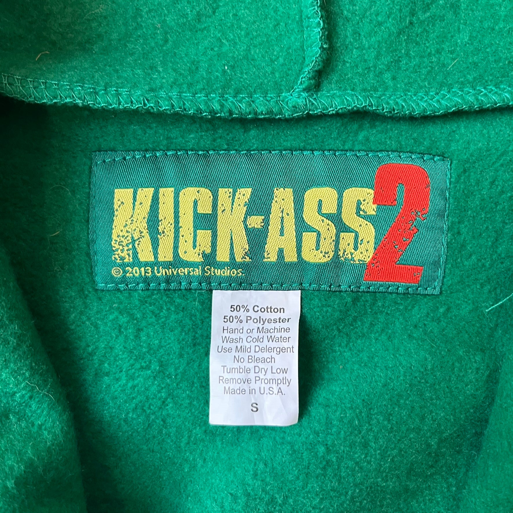 Kick-Ass 2 Full Zip 2013 Promo Hoodie is a green full-zip hoodie with yellow printed lines matching the costume worn by Kick-Ass in Kick-Ass 2. The zipper extends through the hood, allowing it to close into a mask with see-through mesh eye panels. It features a Kick-Ass 2 (2013) Universal Studios licensed neck tag, a matching movie logo patch on the sleeve, and is made in the USA.

