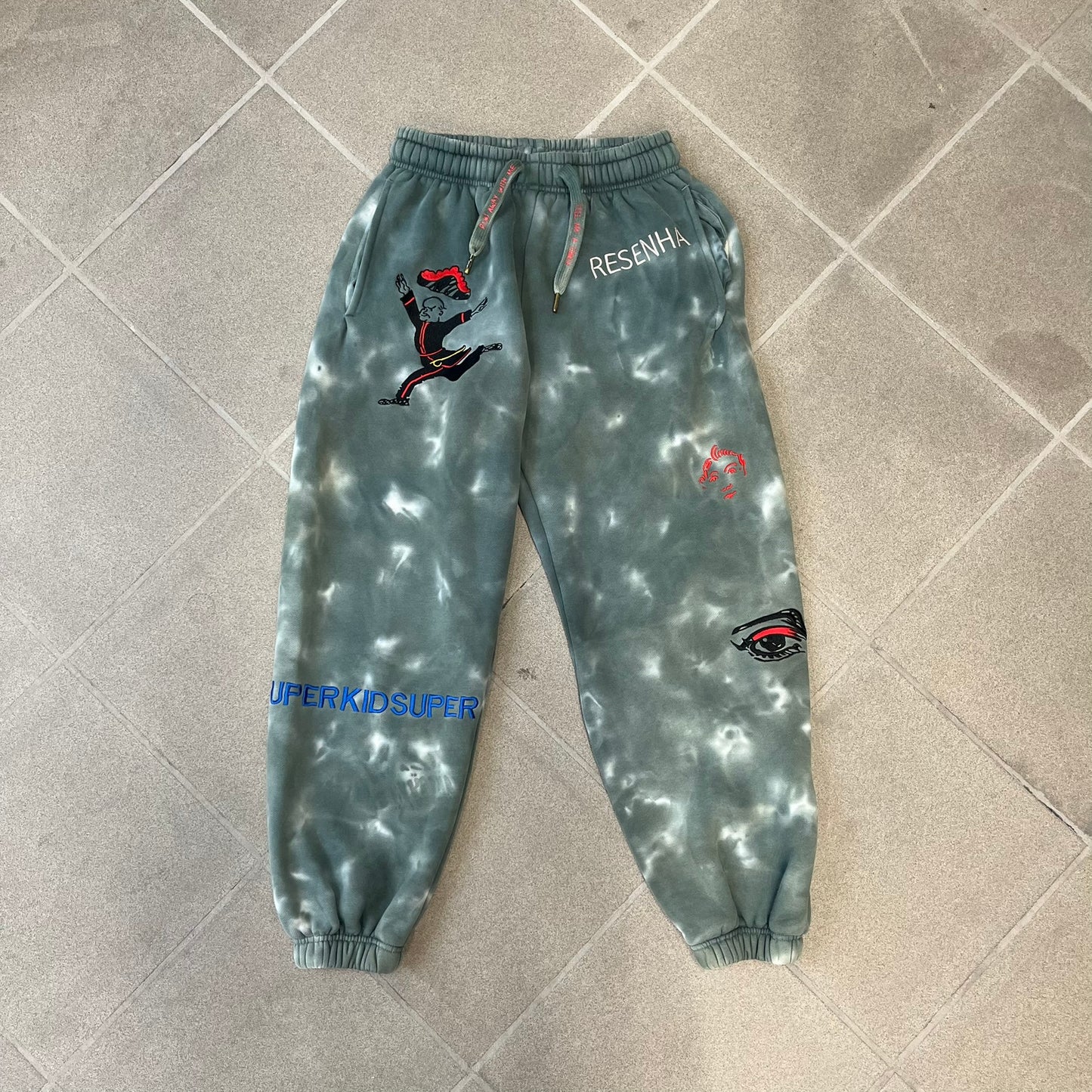 KidSuper Studios FW21 Tie-Dye Sweatpants feature a white and grey tie-dye colorway with multiple embroidered details throughout. The sweatpants include a blue KidSuper logo, eye graphics, “Resenha” text, text on the drawstrings, and additional embroidery on the back pockets. Finished with a pink size tag.