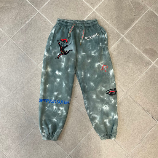KidSuper Studios FW21 Tie-Dye Sweatpants feature a white and grey tie-dye colorway with multiple embroidered details throughout. The sweatpants include a blue KidSuper logo, eye graphics, “Resenha” text, text on the drawstrings, and additional embroidery on the back pockets. Finished with a pink size tag.