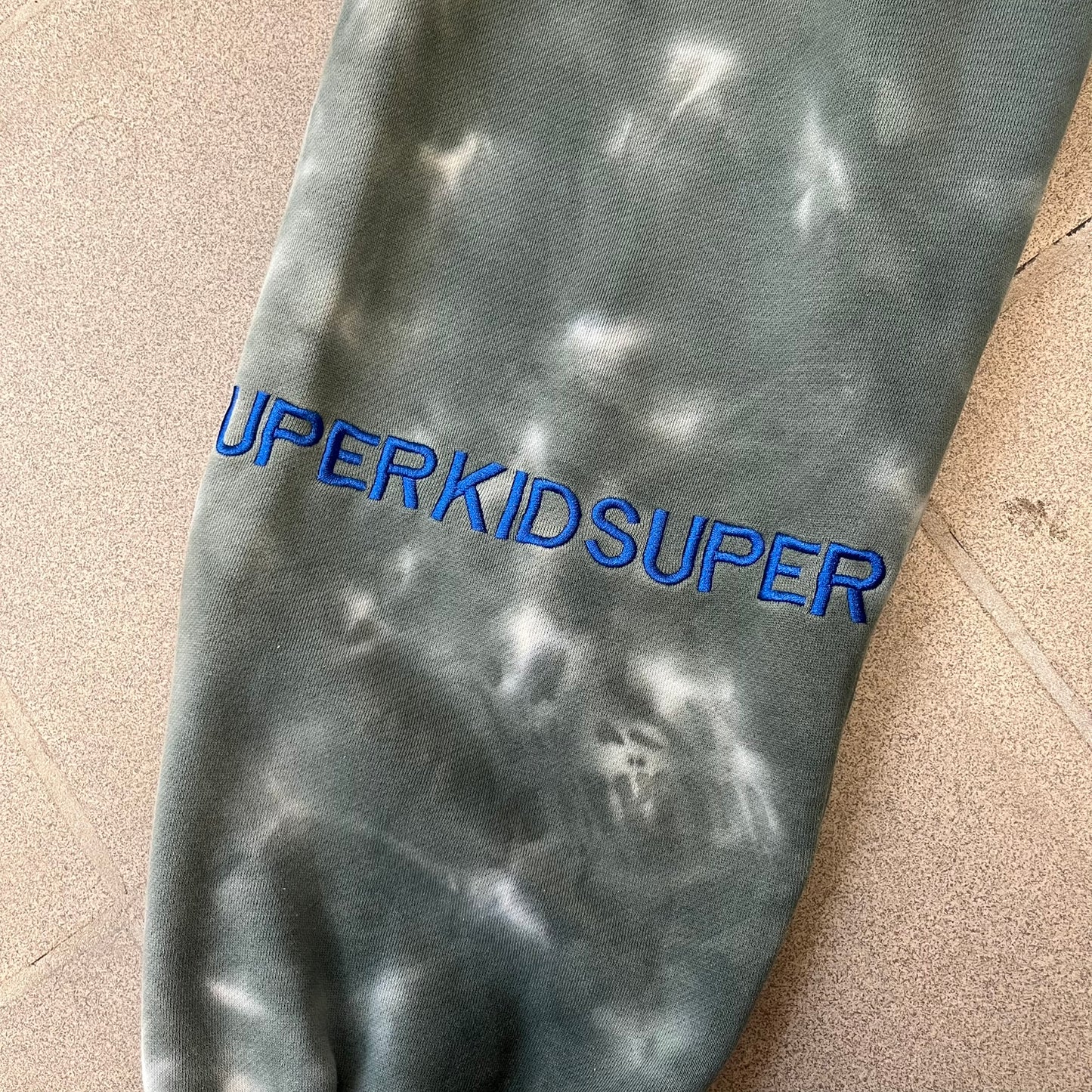 KidSuper Studios FW21 Tie-Dye Sweatpants feature a white and grey tie-dye colorway with multiple embroidered details throughout. The sweatpants include a blue KidSuper logo, eye graphics, “Resenha” text, text on the drawstrings, and additional embroidery on the back pockets. Finished with a pink size tag.