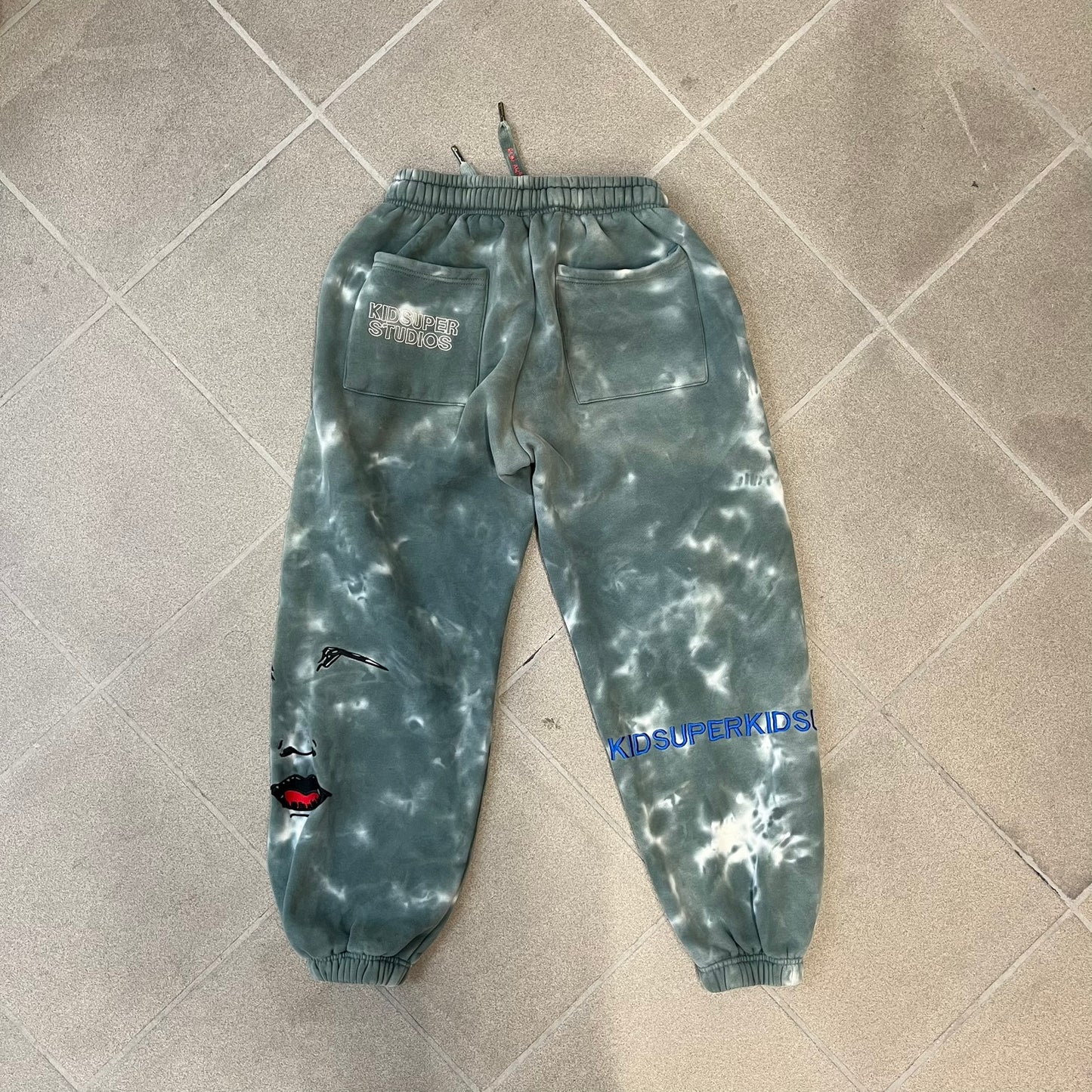 KidSuper Studios FW21 Tie-Dye Sweatpants feature a white and grey tie-dye colorway with multiple embroidered details throughout. The sweatpants include a blue KidSuper logo, eye graphics, “Resenha” text, text on the drawstrings, and additional embroidery on the back pockets. Finished with a pink size tag.