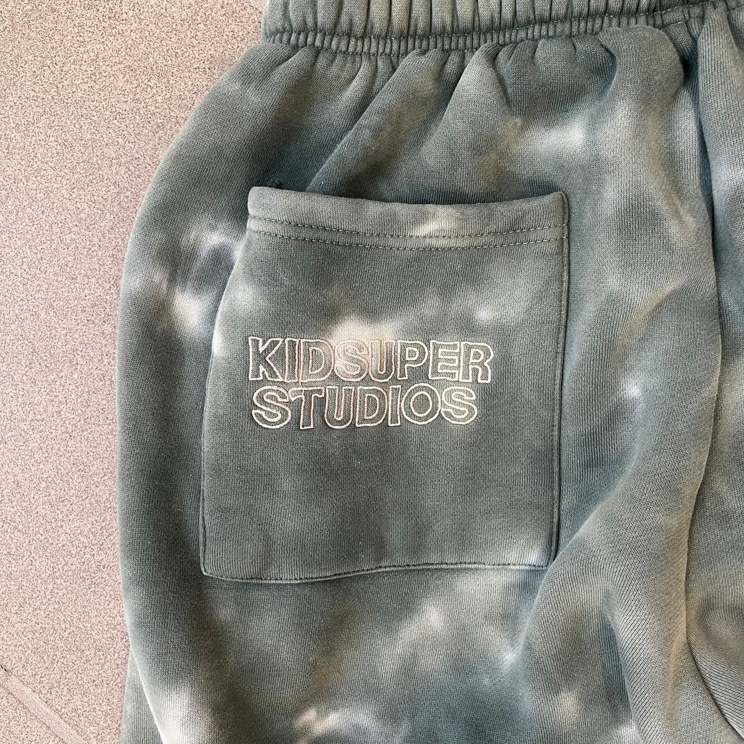 KidSuper Studios FW21 Tie-Dye Sweatpants feature a white and grey tie-dye colorway with multiple embroidered details throughout. The sweatpants include a blue KidSuper logo, eye graphics, “Resenha” text, text on the drawstrings, and additional embroidery on the back pockets. Finished with a pink size tag.