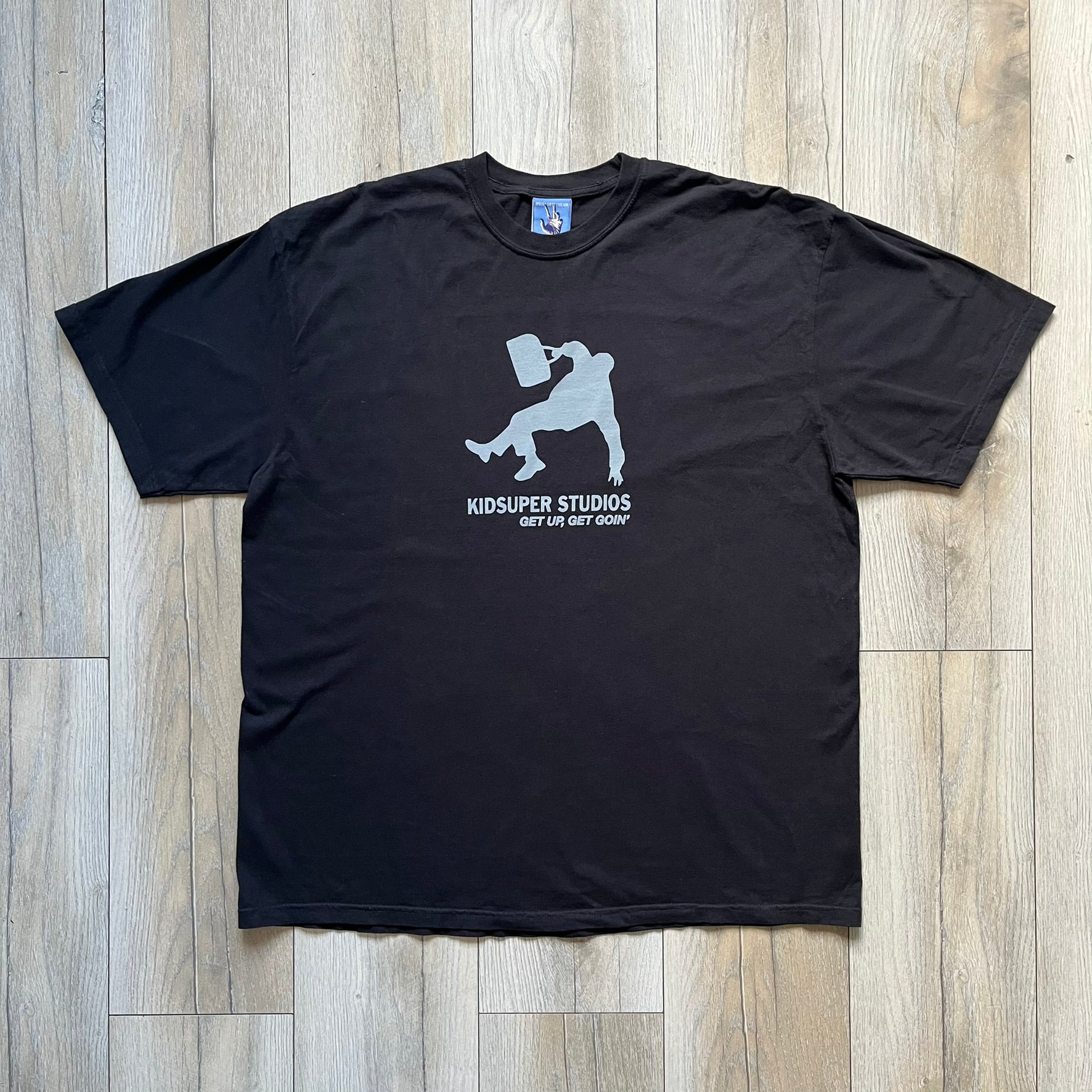 KidSuper Studios SS25 “Get Up, Get Goin’” Black Tee is a black short-sleeve T-shirt from the Spring/Summer 2025 collection. The front features a white and light blue graphic of a figure holding a briefcase mid-jump in a parkour-style pose above the “Kidsuper Studios Get Up, Get Goin’” text. The shirt includes a KidSuper SS25 blue neck tag.
