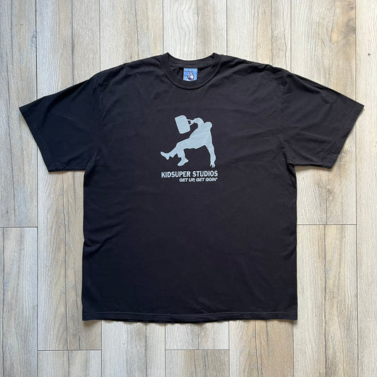 KidSuper Studios SS25 “Get Up, Get Goin’” Black Tee is a black short-sleeve T-shirt from the Spring/Summer 2025 collection. The front features a white and light blue graphic of a figure holding a briefcase mid-jump in a parkour-style pose above the “Kidsuper Studios Get Up, Get Goin’” text. The shirt includes a KidSuper SS25 blue neck tag.
