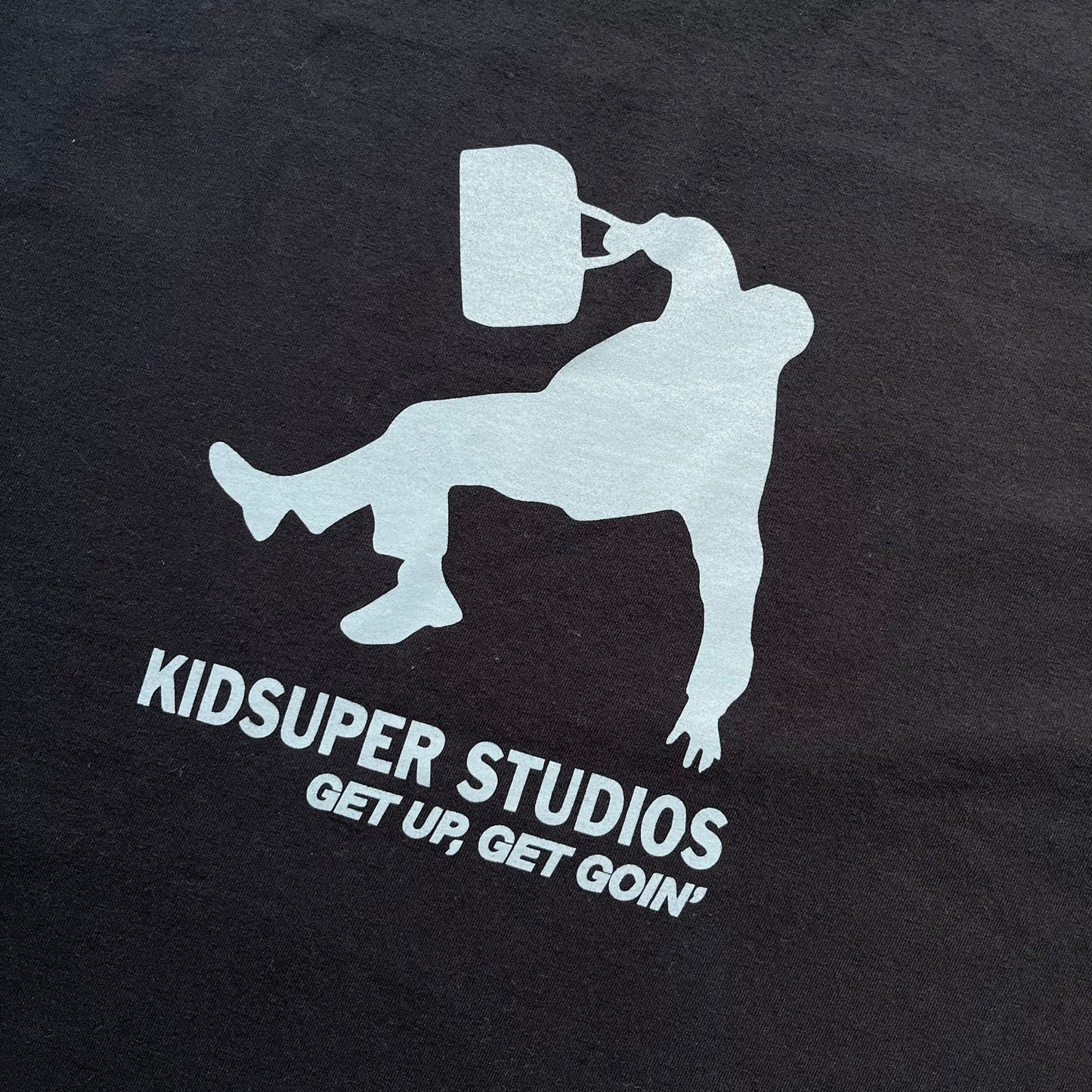 KidSuper Studios SS25 “Get Up, Get Goin’” Black Tee is a black short-sleeve T-shirt from the Spring/Summer 2025 collection. The front features a white and light blue graphic of a figure holding a briefcase mid-jump in a parkour-style pose above the “Kidsuper Studios Get Up, Get Goin’” text. The shirt includes a KidSuper SS25 blue neck tag.
