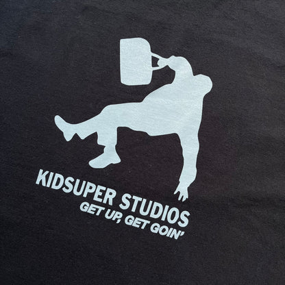 KidSuper Studios SS25 “Get Up, Get Goin’” Black Tee is a black short-sleeve T-shirt from the Spring/Summer 2025 collection. The front features a white and light blue graphic of a figure holding a briefcase mid-jump in a parkour-style pose above the “Kidsuper Studios Get Up, Get Goin’” text. The shirt includes a KidSuper SS25 blue neck tag.
