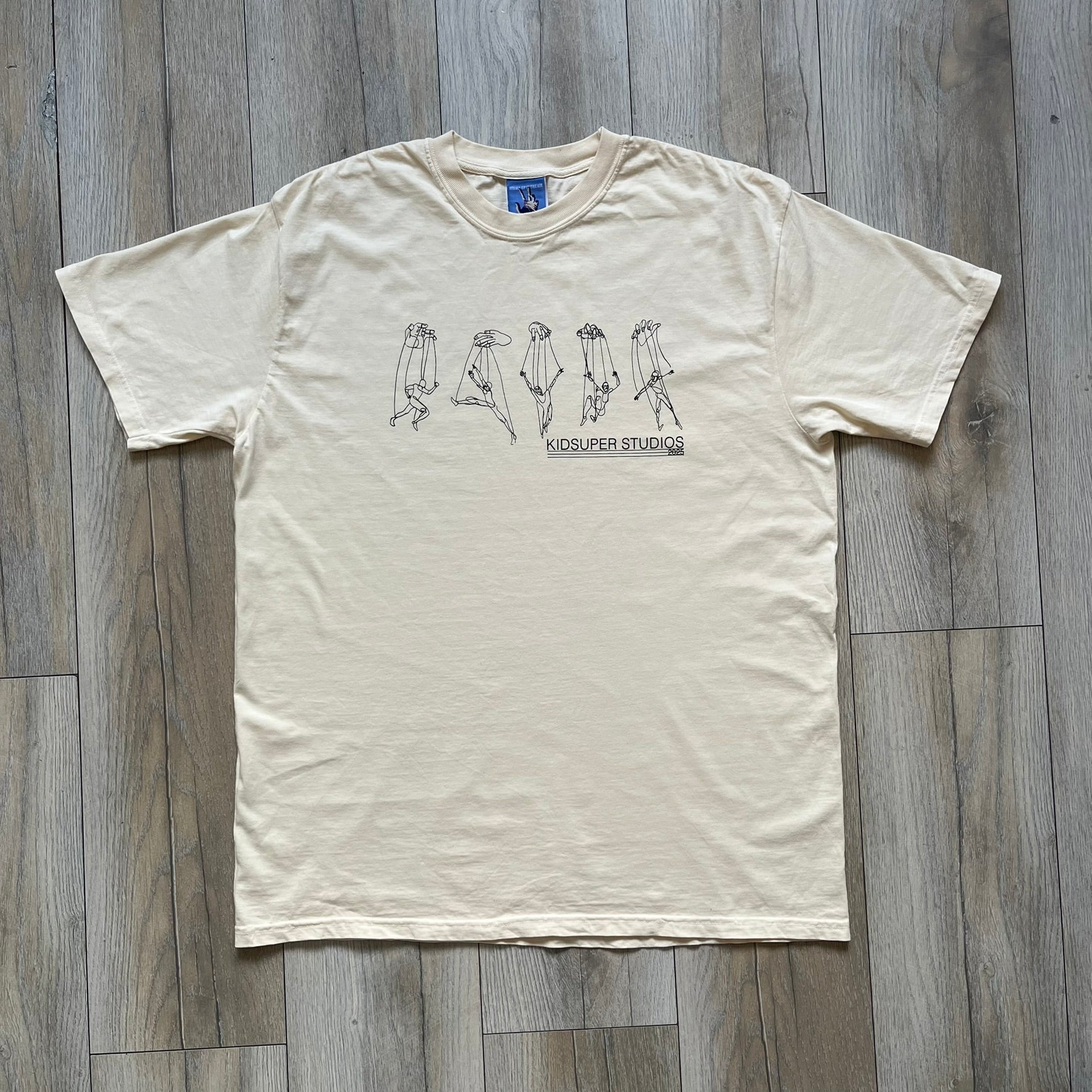 KidSuper Studios SS25 “String Puppets” Cream Tee is a cream short-sleeve T-shirt from the Spring/Summer 2025 collection. The front features five black string puppet-style figures on the chest, with “Kidsuper Studios 2025” text/logo printed underneath. The shirt includes a blue SS25 KidSuper neck tag.