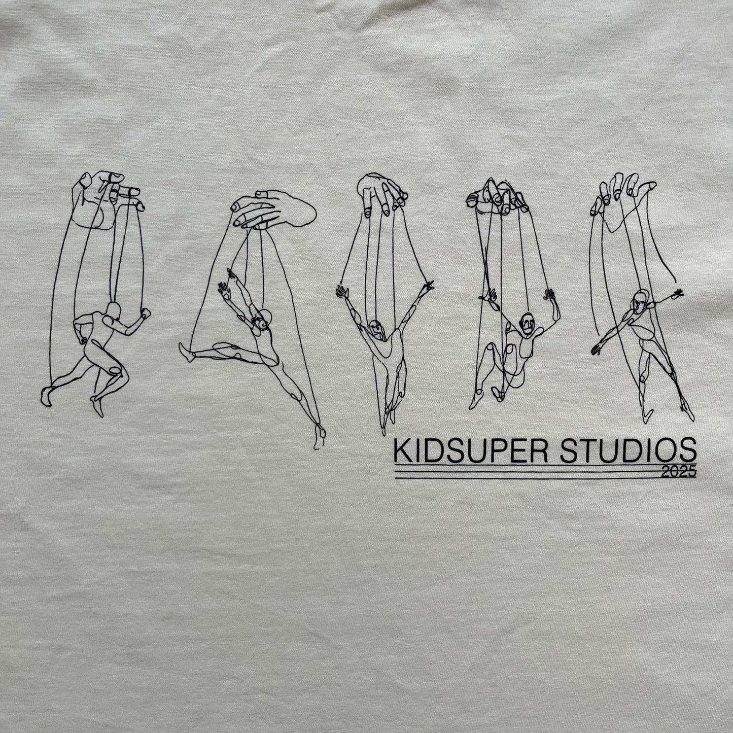 KidSuper Studios SS25 “String Puppets” Cream Tee is a cream short-sleeve T-shirt from the Spring/Summer 2025 collection. The front features five black string puppet-style figures on the chest, with “Kidsuper Studios 2025” text/logo printed underneath. The shirt includes a blue SS25 KidSuper neck tag.