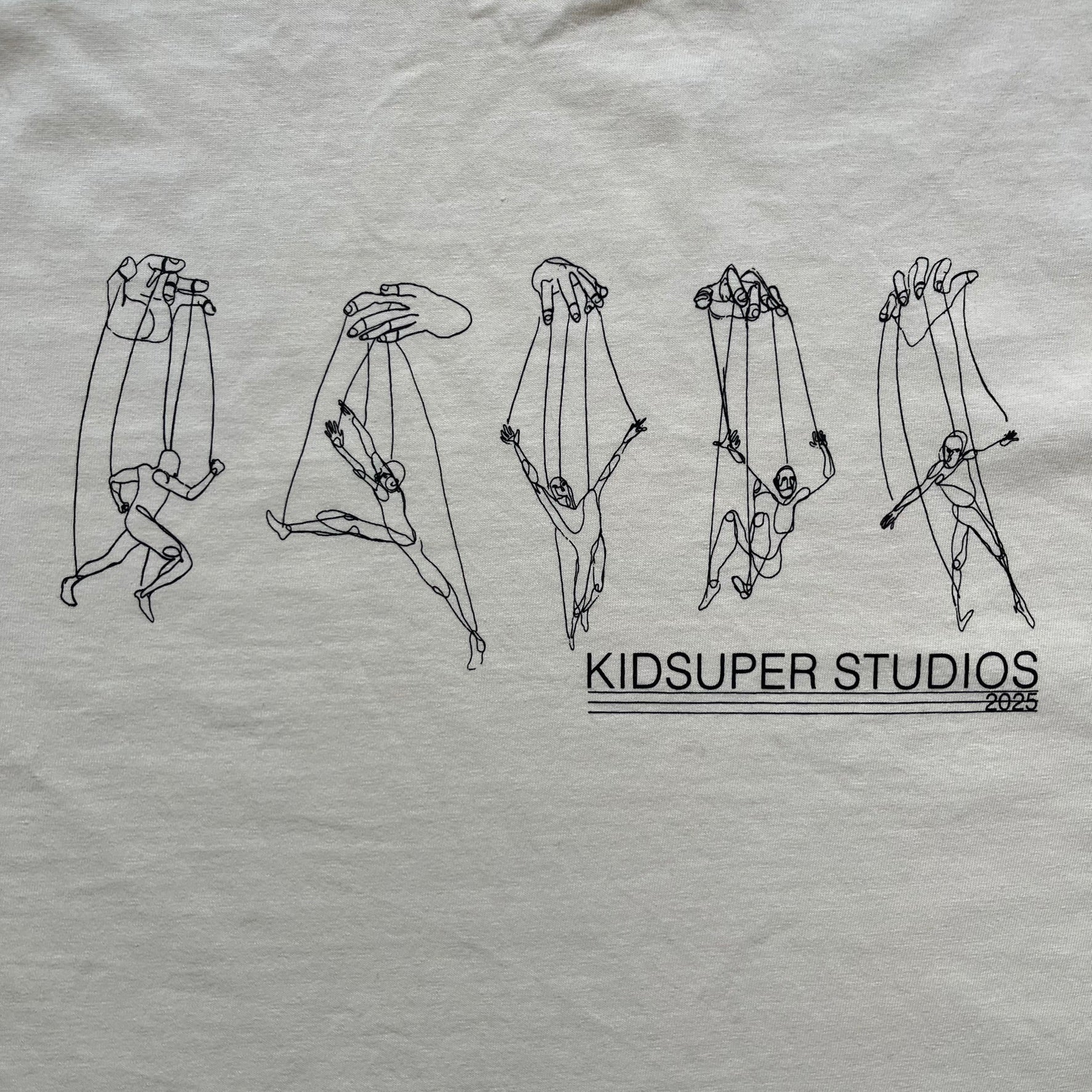 KidSuper Studios SS25 “String Puppets” Cream Tee is a cream short-sleeve T-shirt from the Spring/Summer 2025 collection. The front features five black string puppet-style figures on the chest, with “Kidsuper Studios 2025” text/logo printed underneath. The shirt includes a blue SS25 KidSuper neck tag.