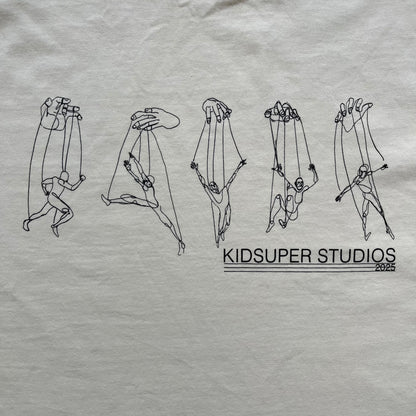 KidSuper Studios SS25 “String Puppets” Cream Tee is a cream short-sleeve T-shirt from the Spring/Summer 2025 collection. The front features five black string puppet-style figures on the chest, with “Kidsuper Studios 2025” text/logo printed underneath. The shirt includes a blue SS25 KidSuper neck tag.