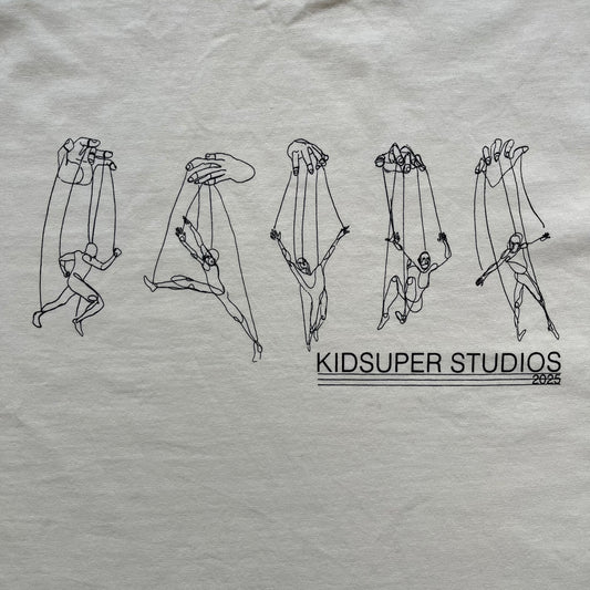 KidSuper Studios SS25 “String Puppets” Cream Tee is a cream short-sleeve T-shirt from the Spring/Summer 2025 collection. The front features five black string puppet-style figures on the chest, with “Kidsuper Studios 2025” text/logo printed underneath. The shirt includes a blue SS25 KidSuper neck tag.