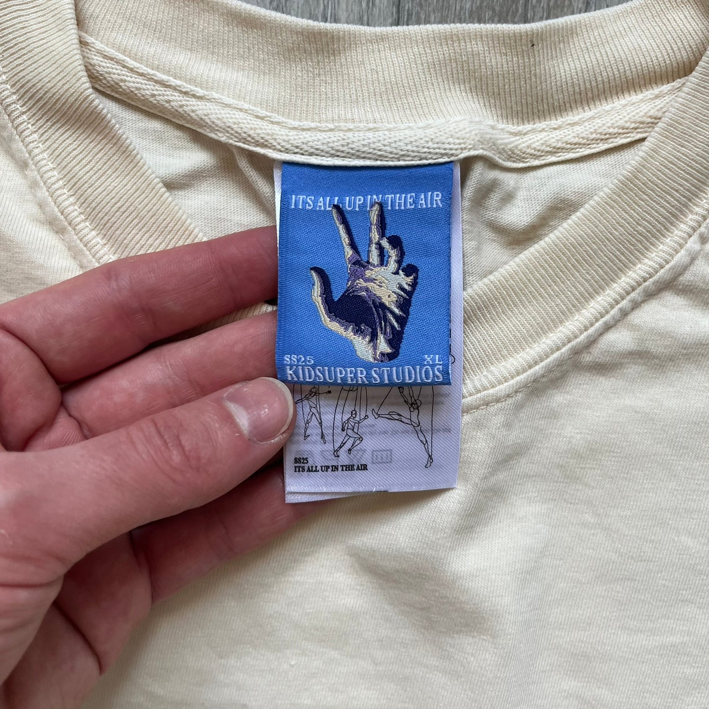 KidSuper Studios SS25 “String Puppets” Cream Tee is a cream short-sleeve T-shirt from the Spring/Summer 2025 collection. The front features five black string puppet-style figures on the chest, with “Kidsuper Studios 2025” text/logo printed underneath. The shirt includes a blue SS25 KidSuper neck tag.