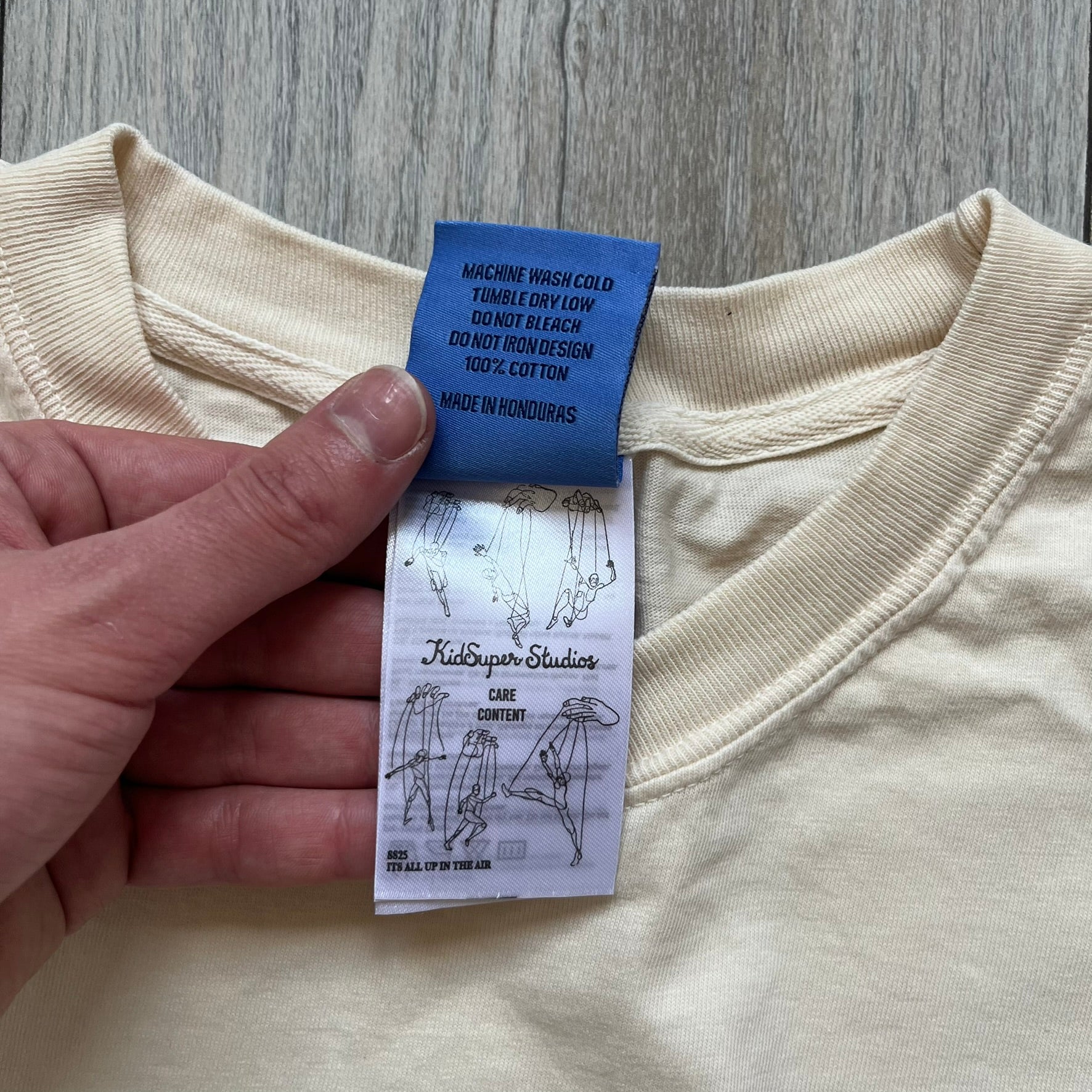 KidSuper Studios SS25 “String Puppets” Cream Tee is a cream short-sleeve T-shirt from the Spring/Summer 2025 collection. The front features five black string puppet-style figures on the chest, with “Kidsuper Studios 2025” text/logo printed underneath. The shirt includes a blue SS25 KidSuper neck tag.