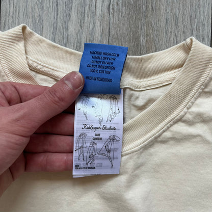KidSuper Studios SS25 “String Puppets” Cream Tee is a cream short-sleeve T-shirt from the Spring/Summer 2025 collection. The front features five black string puppet-style figures on the chest, with “Kidsuper Studios 2025” text/logo printed underneath. The shirt includes a blue SS25 KidSuper neck tag.