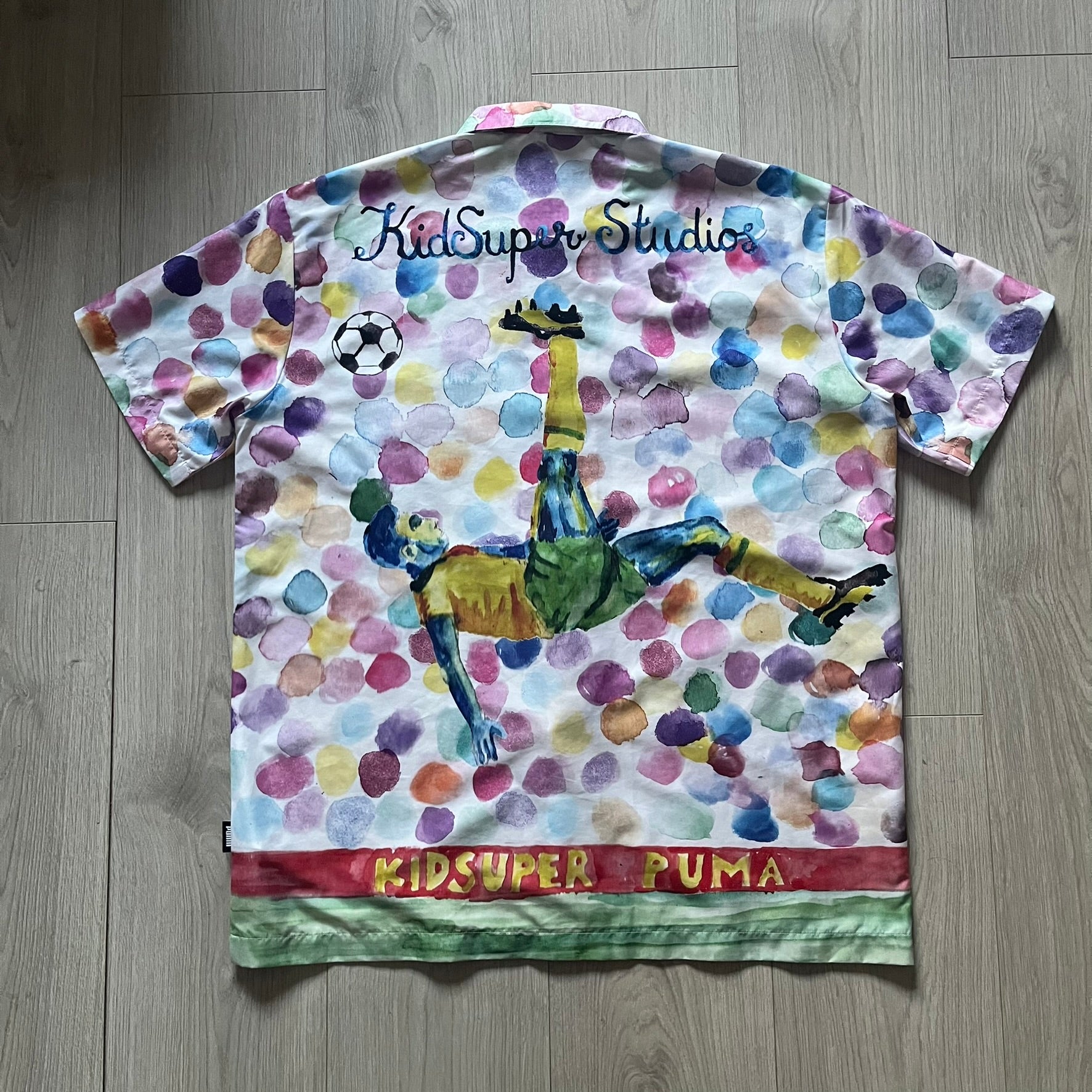 From the KidSuper Studios x Puma 2020 collection, this reversible football shirt (598953-02) showcases a watercolor-style all-over crowd print with vibrant color detailing. The design includes a red and yellow co-branded banner, a green field stripe and a back graphic of a football player doing a bicycle kick. Complete with a chest pocket and embroidered KidSuper Studios x Puma logo.