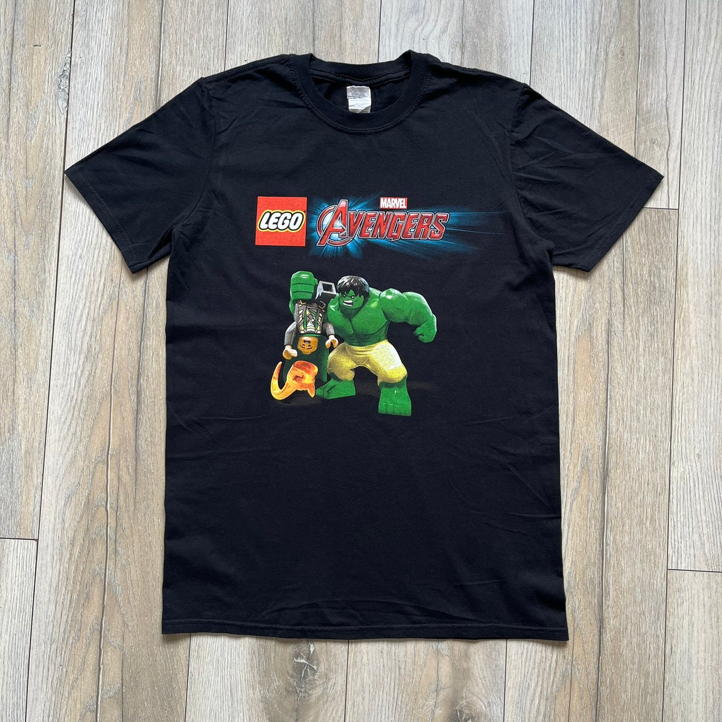 LEGO Avengers 2016 Promo Tee is a black T-shirt featuring a LEGO Marvel Avengers logo on the front. Below the logo is a graphic of LEGO Hulk holding Loki upside down. The shirt is made on a Gildan blank and includes 2016 LEGO and Marvel licensing inside the shirt.
