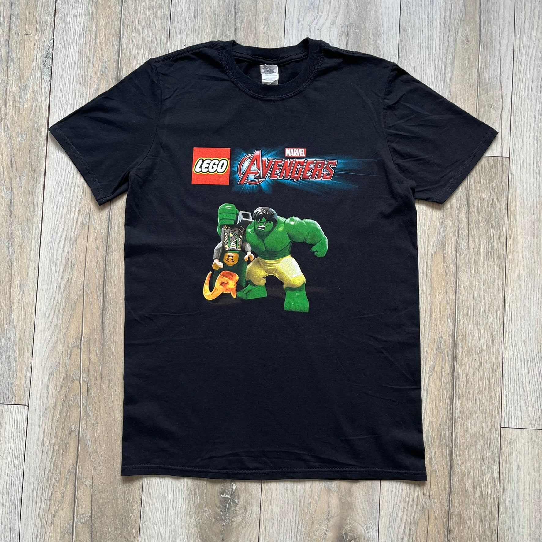 LEGO Avengers 2016 Promo Tee is a black T-shirt featuring a LEGO Marvel Avengers logo on the front. Below the logo is a graphic of LEGO Hulk holding Loki upside down. The shirt is made on a Gildan blank and includes 2016 LEGO and Marvel licensing inside the shirt.