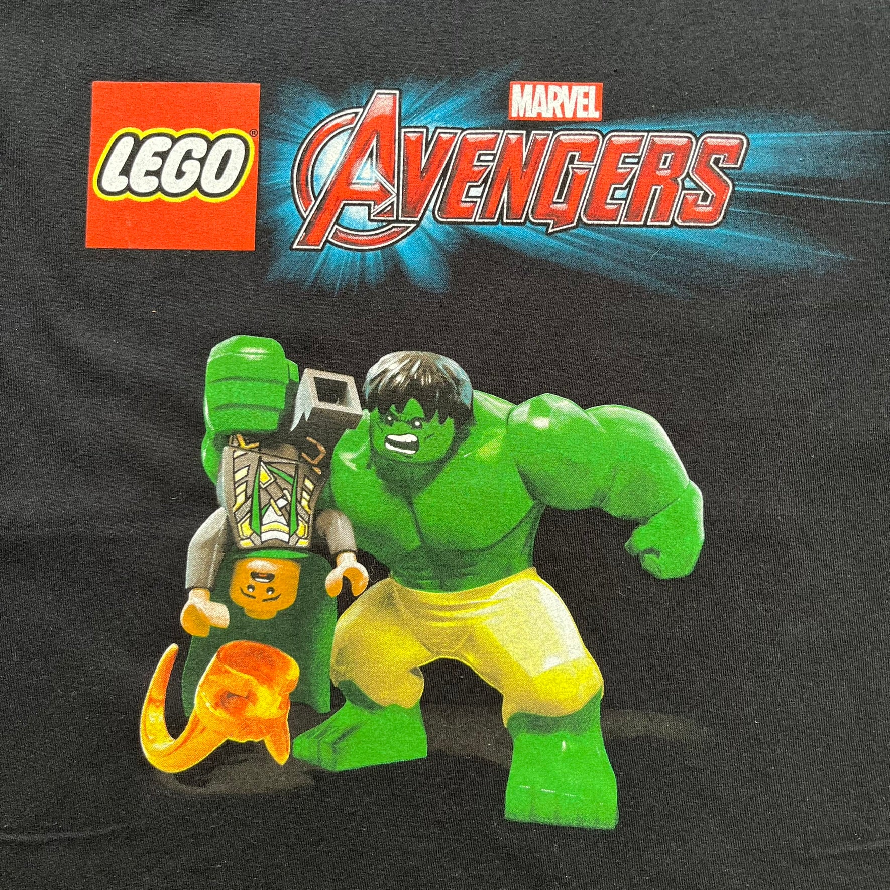 LEGO Avengers 2016 Promo Tee is a black T-shirt featuring a LEGO Marvel Avengers logo on the front. Below the logo is a graphic of LEGO Hulk holding Loki upside down. The shirt is made on a Gildan blank and includes 2016 LEGO and Marvel licensing inside the shirt.