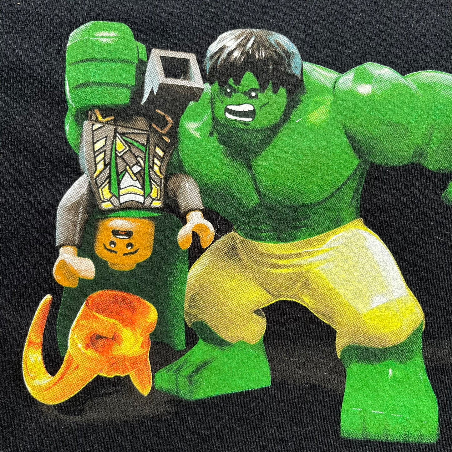 LEGO Avengers 2016 Promo Tee is a black T-shirt featuring a LEGO Marvel Avengers logo on the front. Below the logo is a graphic of LEGO Hulk holding Loki upside down. The shirt is made on a Gildan blank and includes 2016 LEGO and Marvel licensing inside the shirt.