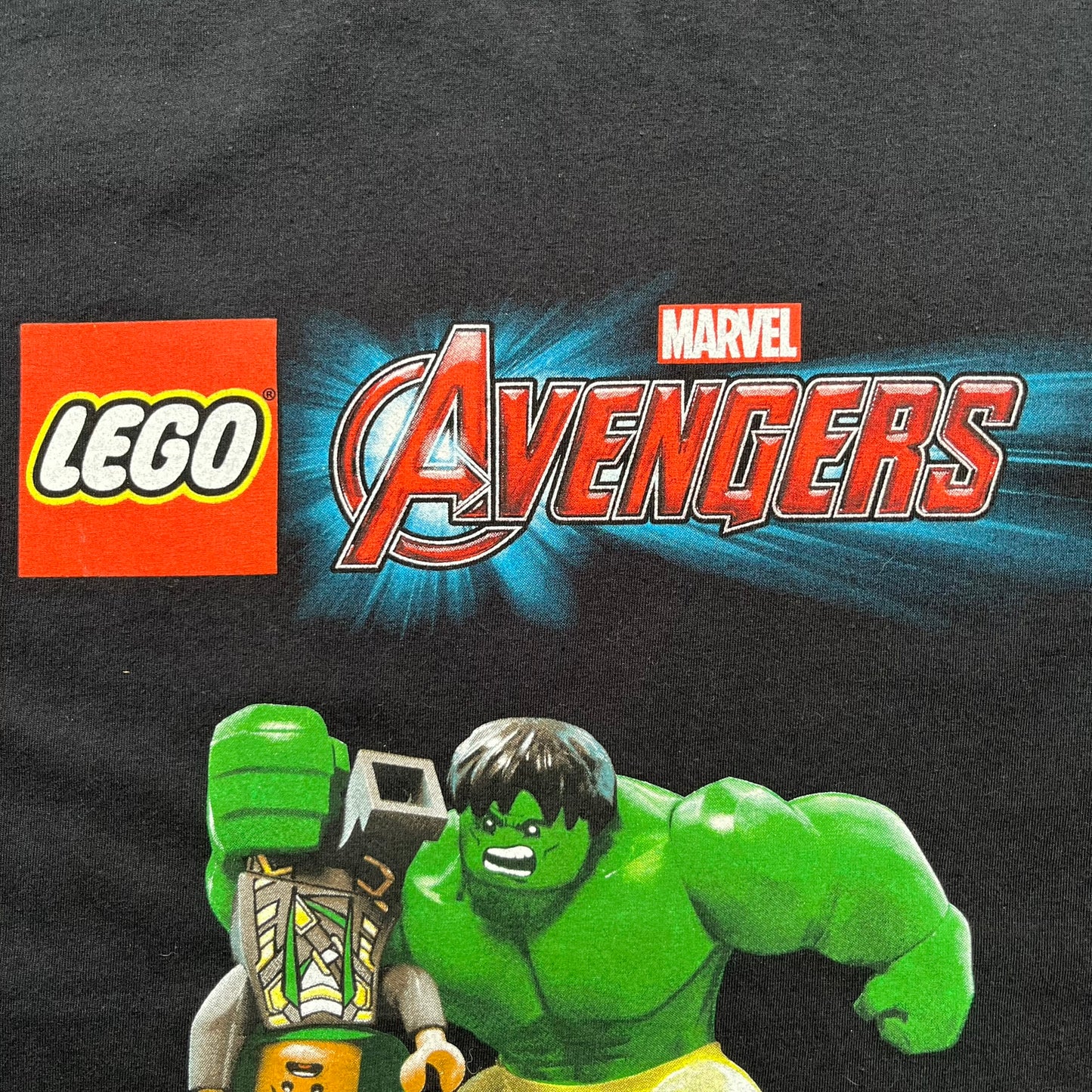 LEGO Avengers 2016 Promo Tee is a black T-shirt featuring a LEGO Marvel Avengers logo on the front. Below the logo is a graphic of LEGO Hulk holding Loki upside down. The shirt is made on a Gildan blank and includes 2016 LEGO and Marvel licensing inside the shirt.