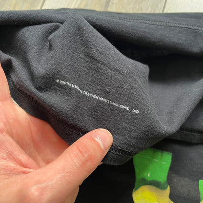 LEGO Avengers 2016 Promo Tee is a black T-shirt featuring a LEGO Marvel Avengers logo on the front. Below the logo is a graphic of LEGO Hulk holding Loki upside down. The shirt is made on a Gildan blank and includes 2016 LEGO and Marvel licensing inside the shirt.