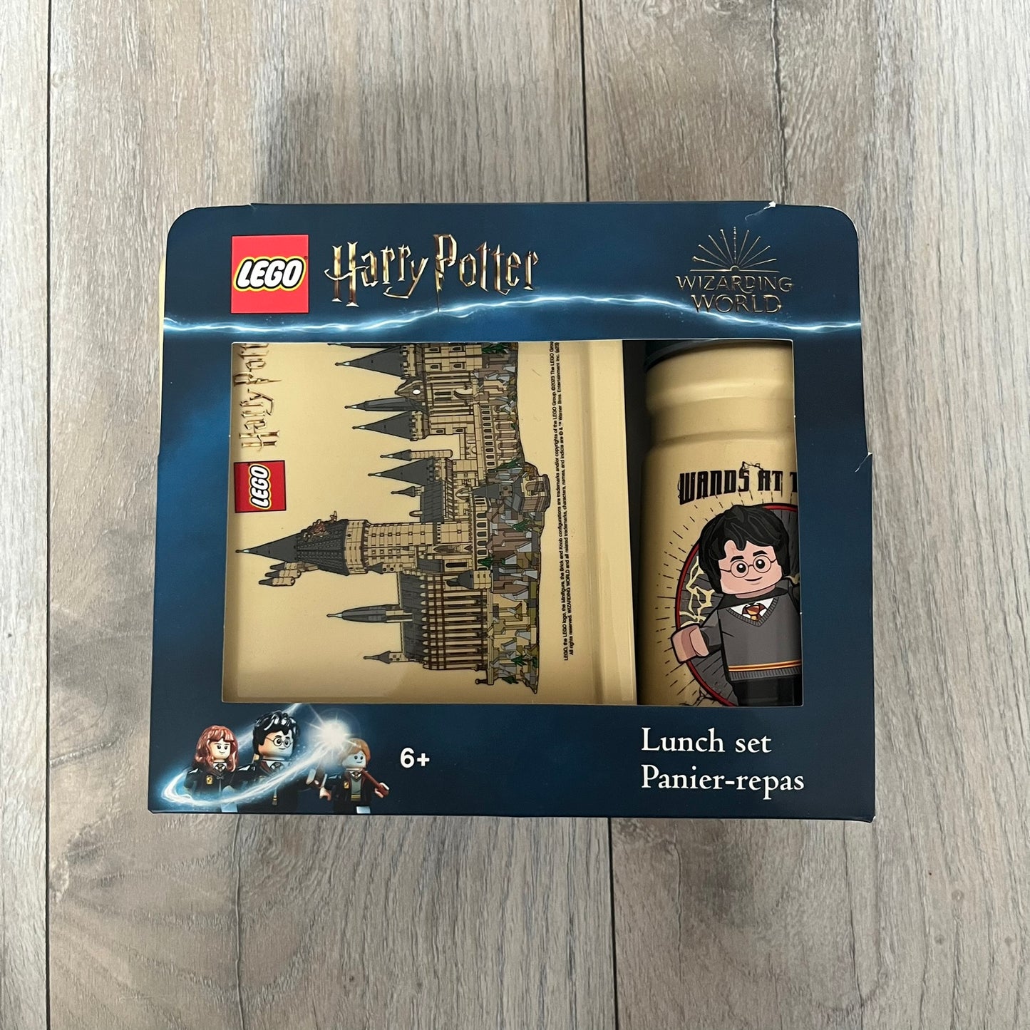 LEGO Harry Potter 2023 Hogwarts Lunch Set is a beige-themed lunch set designed for children aged 3 and up. It includes a polypropylene lunch box and a 390 mL drinking bottle with a silicone spout and hygiene cap. Both items feature illustrations of LEGO Harry Potter characters. The lunch box has easy-open clips, a movable internal divider for separating food, and a lid that can also be used as a plate. The set is designed for portability and everyday use.