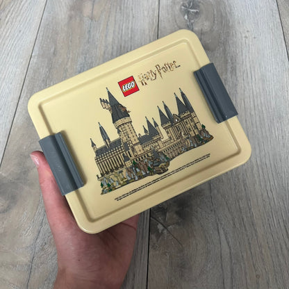 LEGO Harry Potter 2023 Hogwarts Lunch Set is a beige-themed lunch set designed for children aged 3 and up. It includes a polypropylene lunch box and a 390 mL drinking bottle with a silicone spout and hygiene cap. Both items feature illustrations of LEGO Harry Potter characters. The lunch box has easy-open clips, a movable internal divider for separating food, and a lid that can also be used as a plate. The set is designed for portability and everyday use.