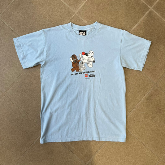 LEGO Star Wars 2007 “Let the WOOKIEE win!” Tee is a light blue, 100% cotton T-shirt with a front graphic of LEGO Chewbacca and two Stormtroopers, with Chewbacca holding the hands of one while the other stands to the side with a blaster. “Let the Wookiee win” text sits below the graphic, along with a small LEGO Star Wars logo and 2007 licensing. It has a Star Wars neck tag.