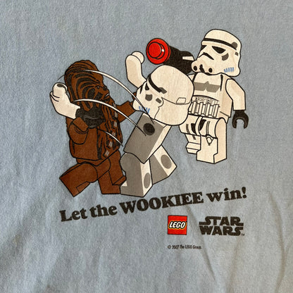LEGO Star Wars 2007 “Let the WOOKIEE win!” Tee is a light blue, 100% cotton T-shirt with a front graphic of LEGO Chewbacca and two Stormtroopers, with Chewbacca holding the hands of one while the other stands to the side with a blaster. “Let the Wookiee win” text sits below the graphic, along with a small LEGO Star Wars logo and 2007 licensing. It has a Star Wars neck tag.