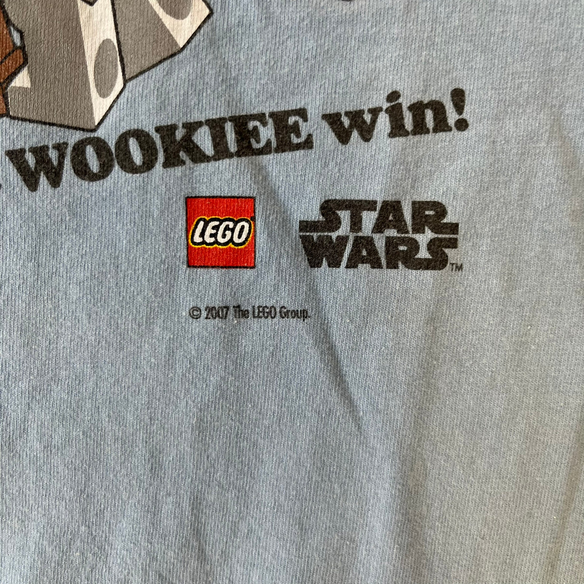LEGO Star Wars 2007 “Let the WOOKIEE win!” Tee is a light blue, 100% cotton T-shirt with a front graphic of LEGO Chewbacca and two Stormtroopers, with Chewbacca holding the hands of one while the other stands to the side with a blaster. “Let the Wookiee win” text sits below the graphic, along with a small LEGO Star Wars logo and 2007 licensing. It has a Star Wars neck tag.